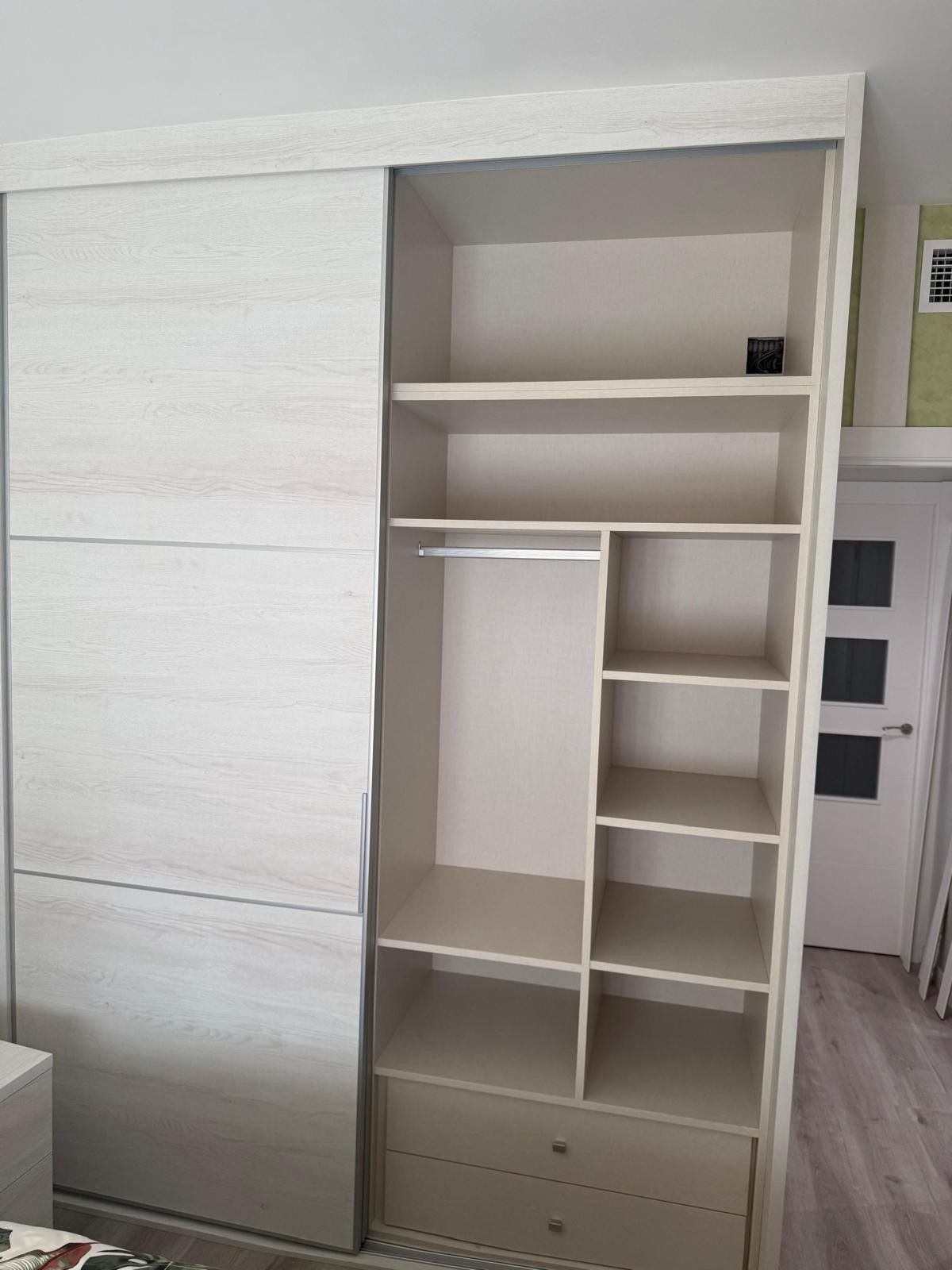 Armario empotrado blanco con puerta corredera, estantes abiertos, espacio para colgar ropa y cajones en un dormitorio.