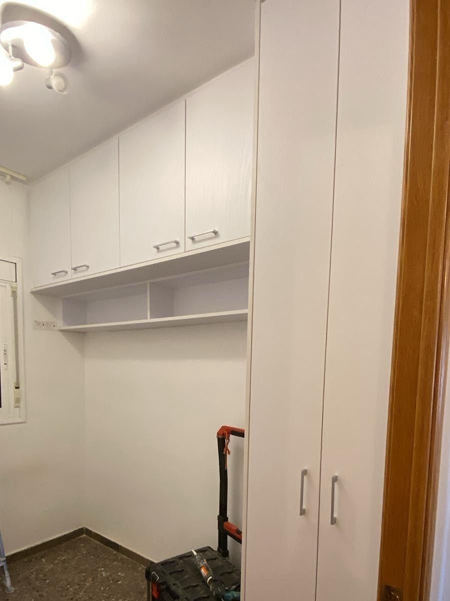 Pequeño cuarto de lavandería blanco con armarios de pared, estante abierto y una aspiradora cerca de la puerta.