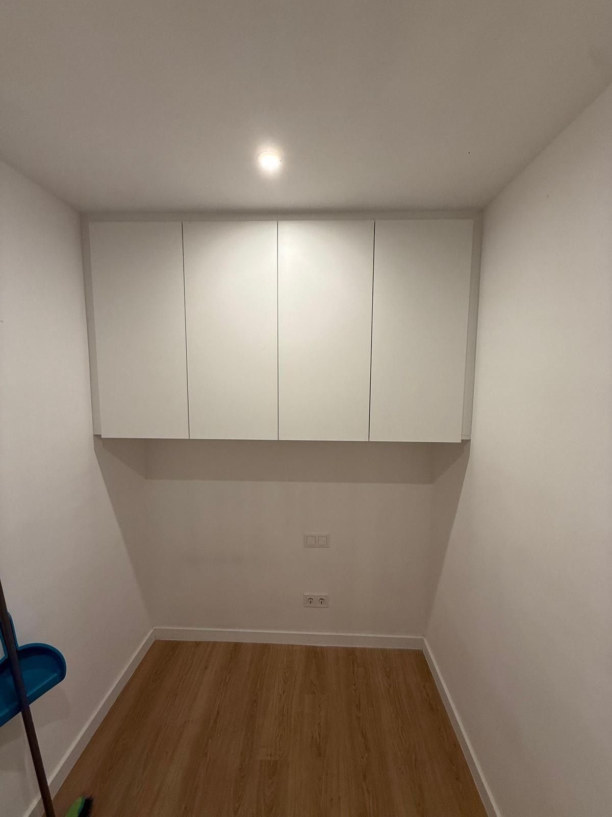 Habitación pequeña y vacía con armarios blancos en la pared del fondo, suelo de madera y una lámpara de techo.