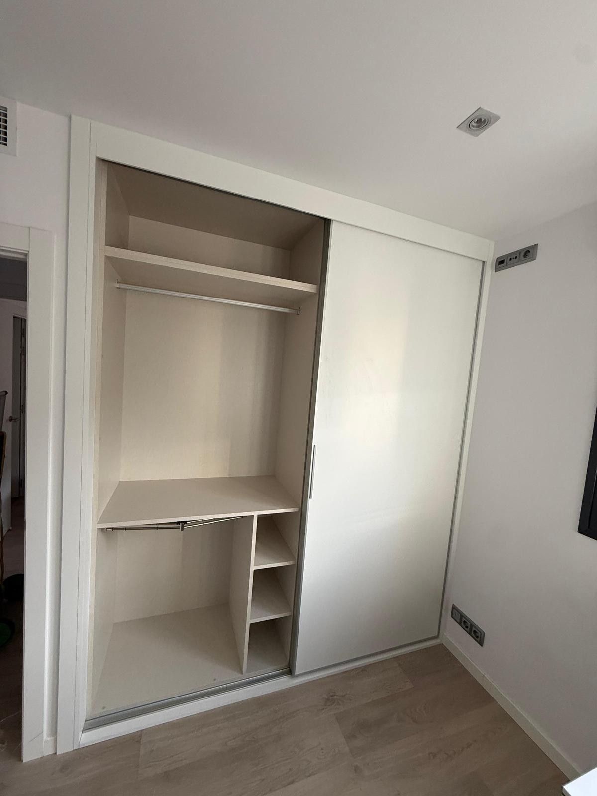 Armario abierto con estantes y barra para colgar ropa en una habitación blanca, con puerta corredera y suelo de madera.