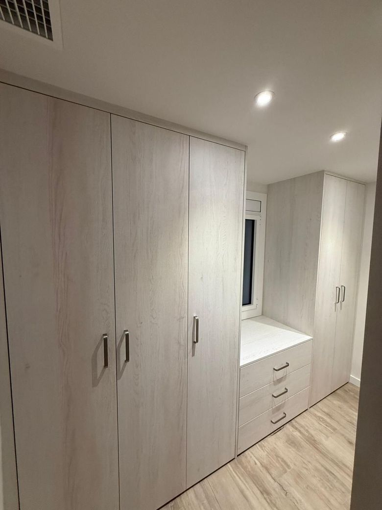 Dormitorio con armarios empotrados blancos con cajones, una ventana y luces empotradas en el techo