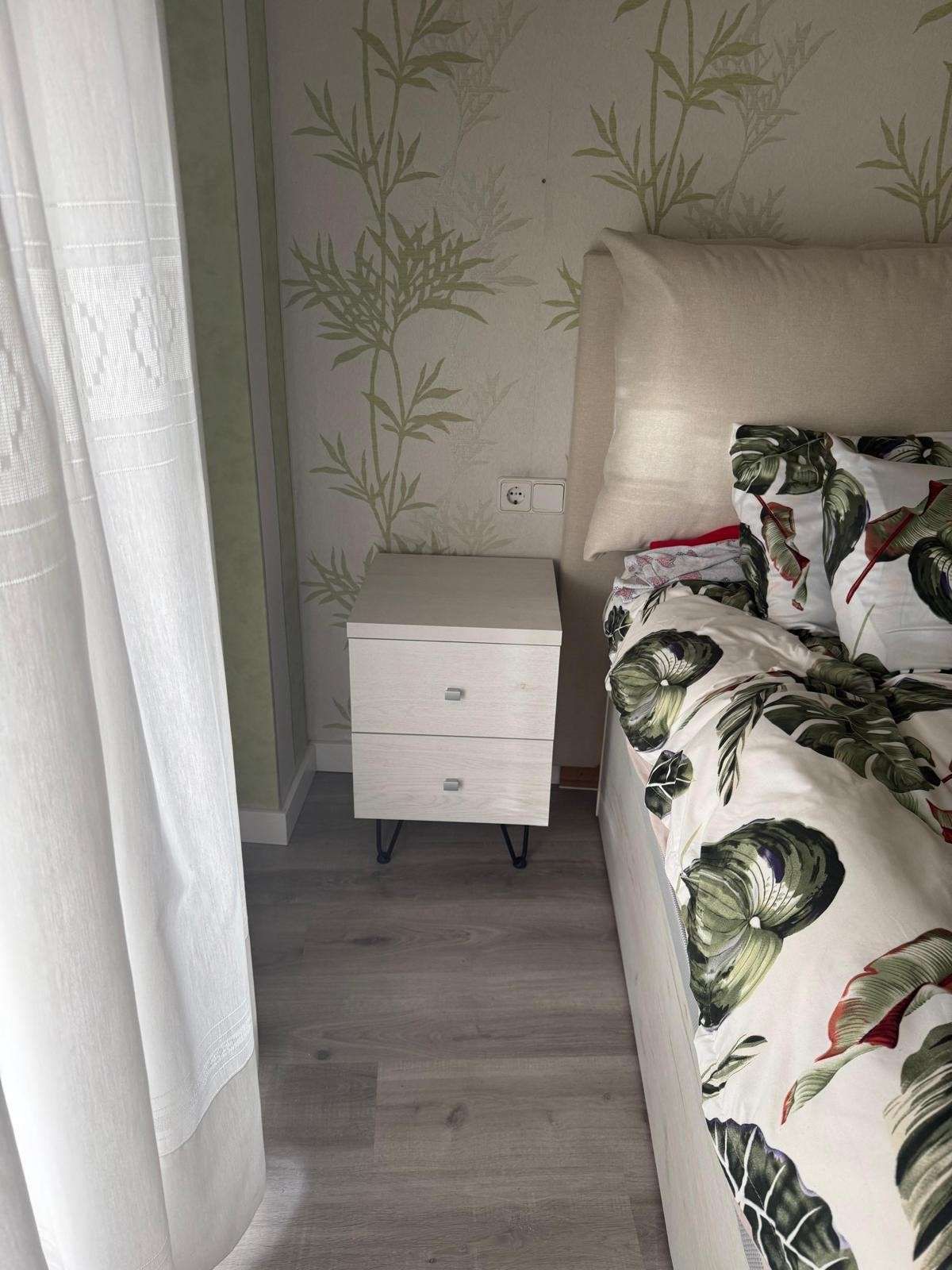 Rincón del dormitorio con mesita de noche blanca junto a la cama, papel pintado de bambú y una cortina transparente.