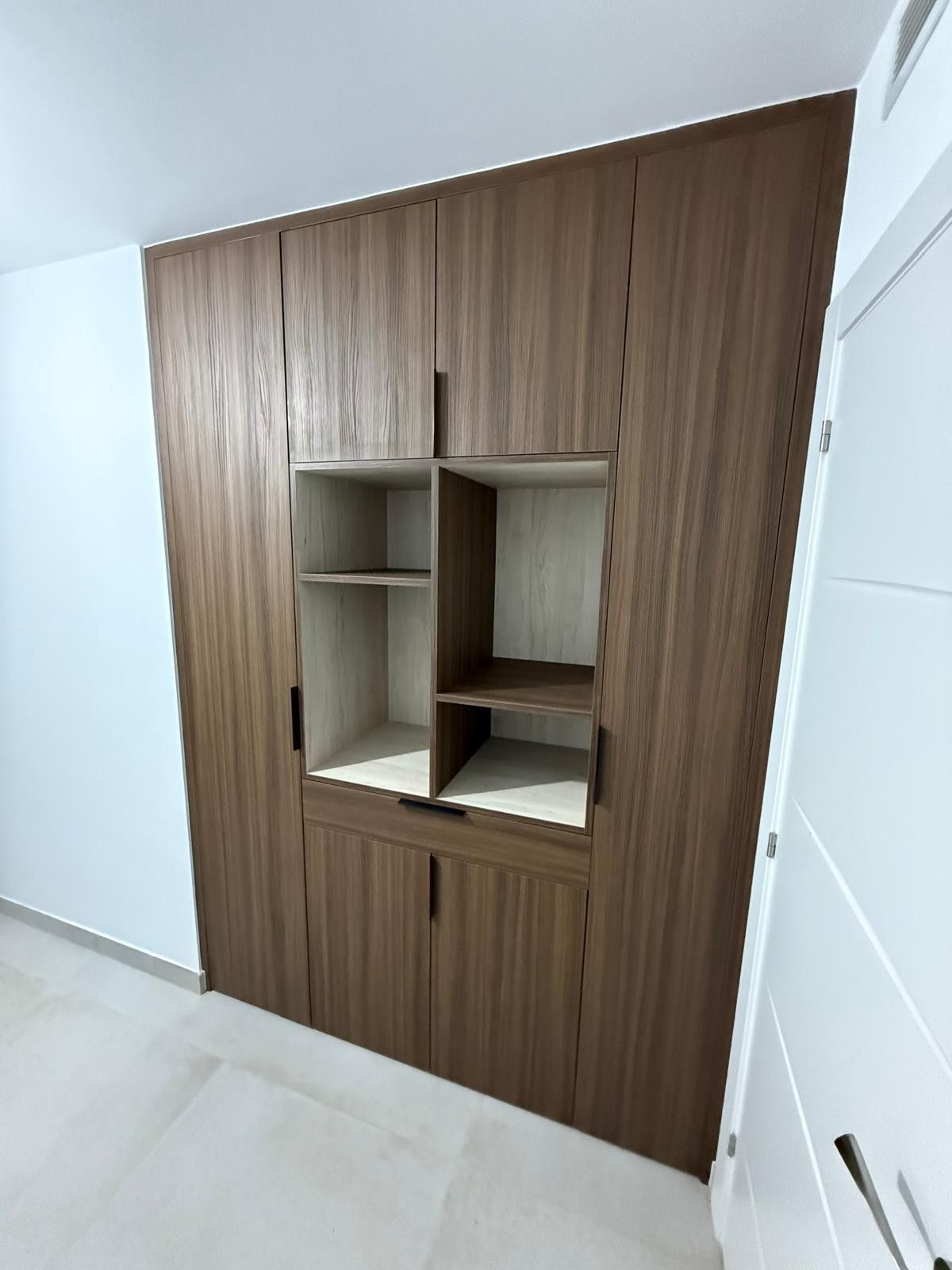 Armario empotrado de madera con estantes abiertos y puerta blanca en un pasillo alicatado.