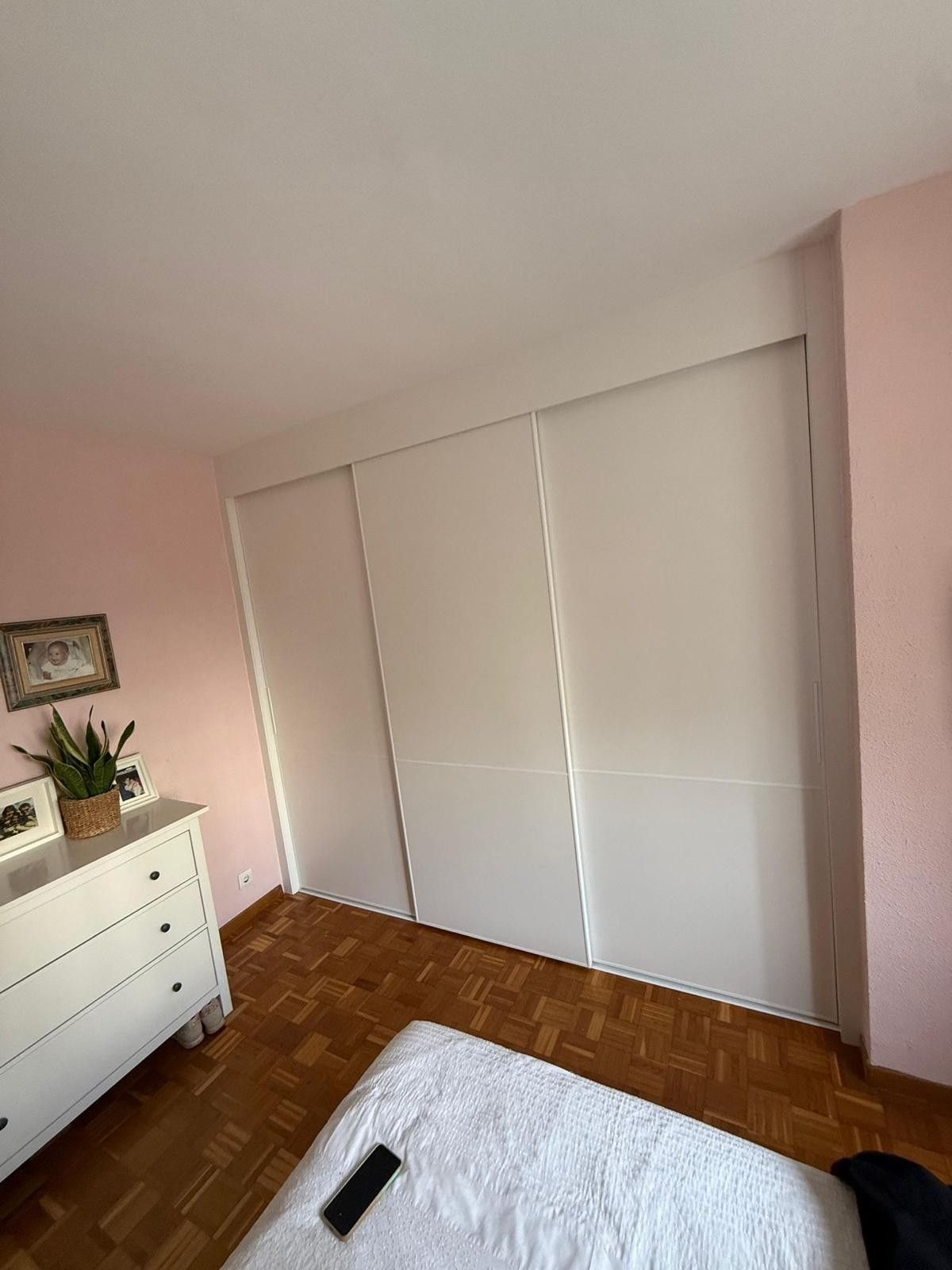 Dormitorio rosa con puertas correderas blancas en el armario, cómoda blanca y una cama en primer plano.