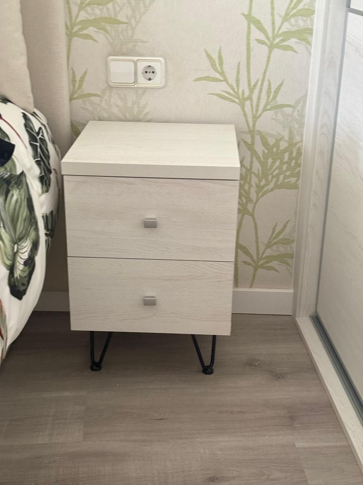 Mesita de noche pequeña, blanca, con dos cajones y patas negras, situada junto a la cama en un dormitorio.