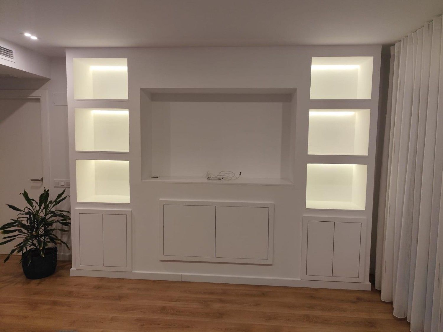 Estanterías y armarios empotrados con iluminación tenue en una habitación moderna, suelo de madera y cortinas blancas.