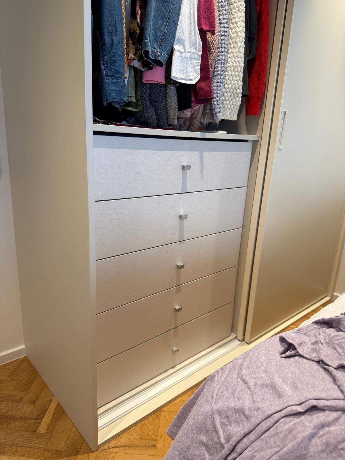 Un armario abierto con ropa colgada sobre cajones blancos y una cama con una manta morada cerca.