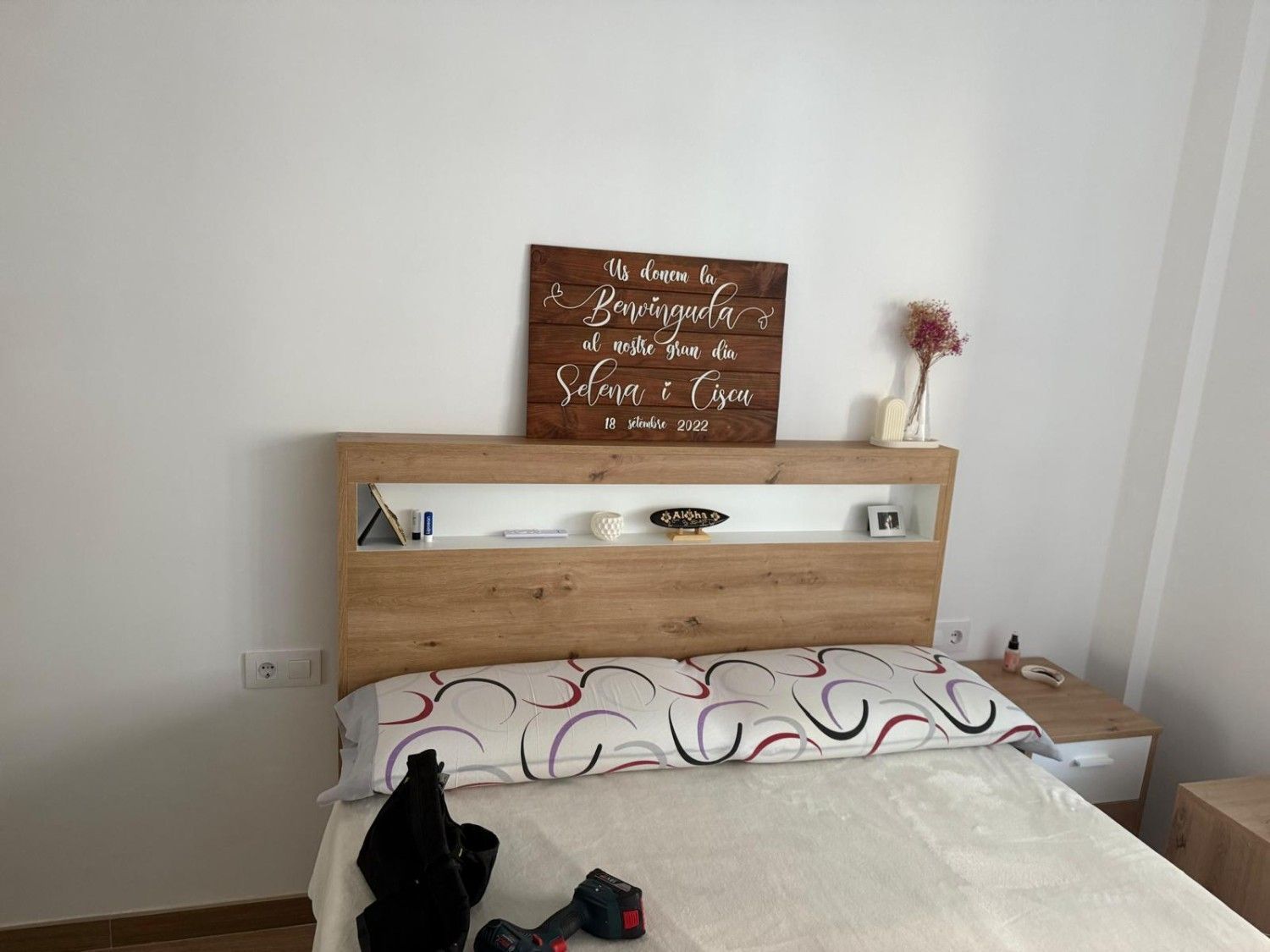 Dormitorio con cama de madera, ropa de cama blanca, cartel en la pared y un jarrón con flores