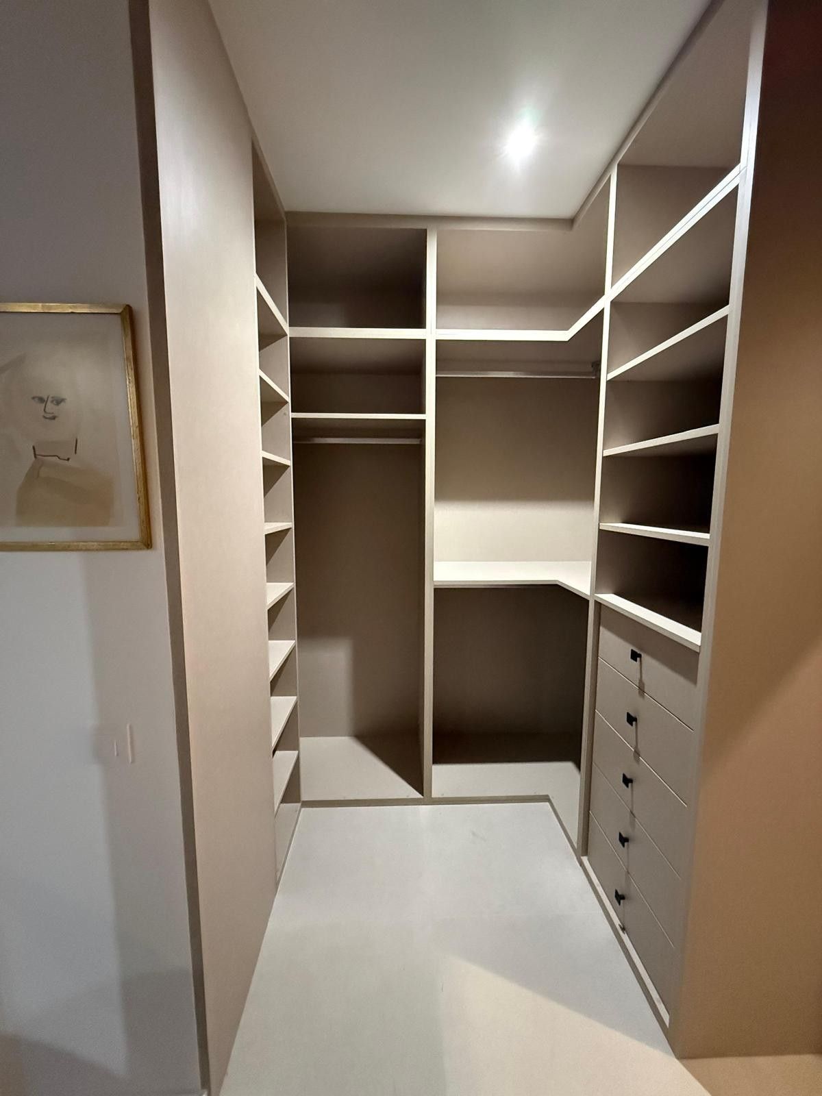 Vestidor con estantes blancos, barras para colgar ropa y cajones bajo una brillante luz de techo.