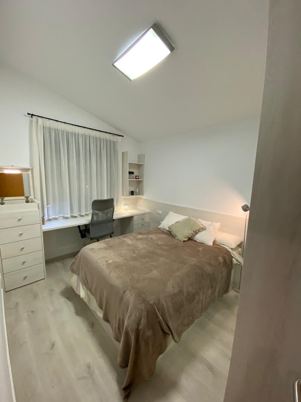 Habitación luminosa con cama, cómoda, escritorio, ventana y luz de techo encendida.