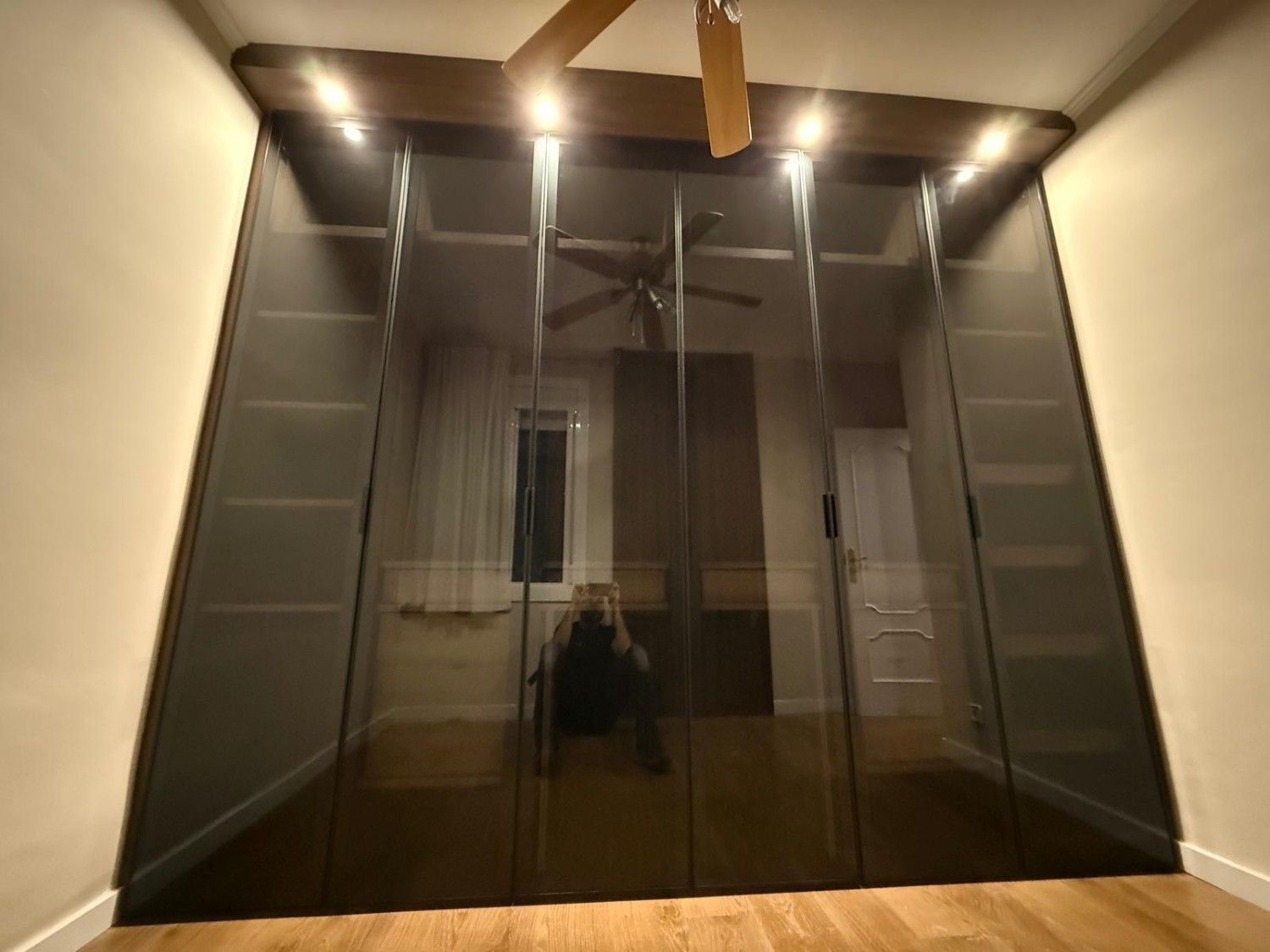 Ascensor moderno de cristal con paneles tintados oscuros en un pasillo interior con iluminación cálida.