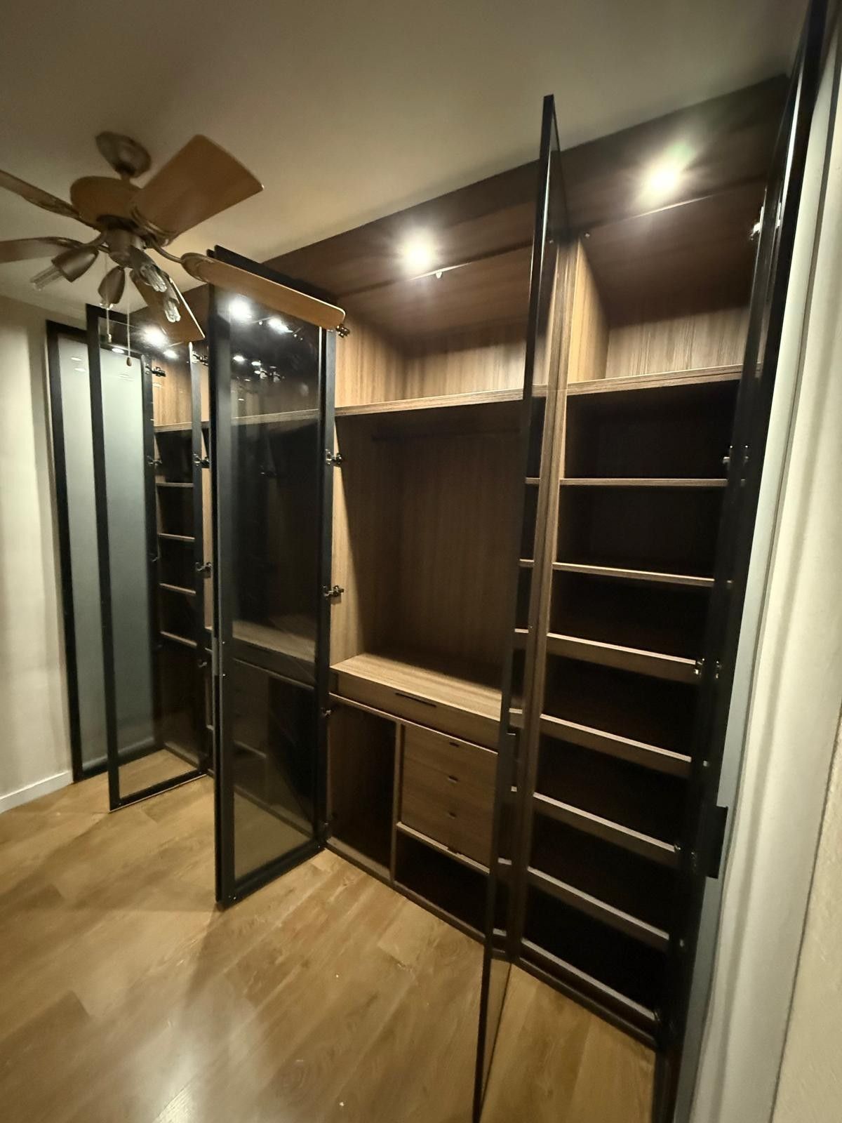 Vestidor con estantes de madera oscura, puertas con espejo y ventilador de techo a la izquierda.