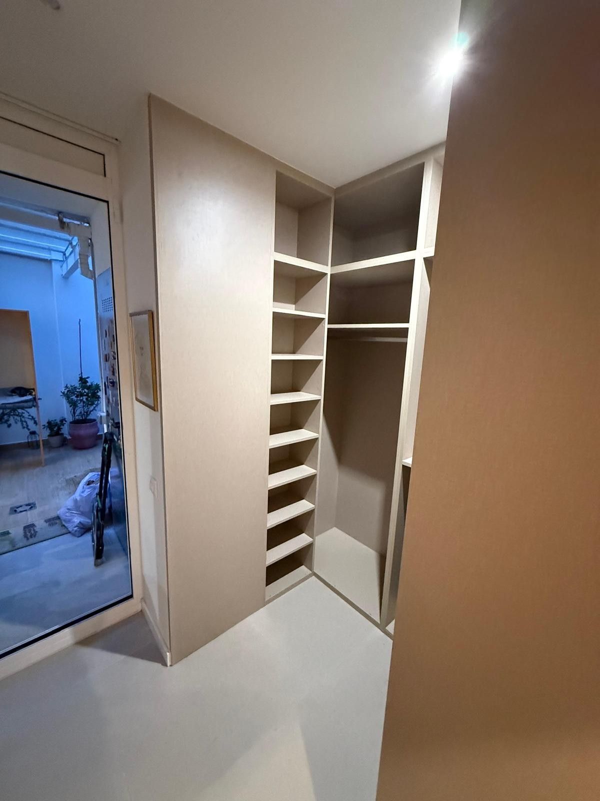 Armario empotrado de color beige con estantes y espacio para colgar ropa, situado junto a una puerta abierta