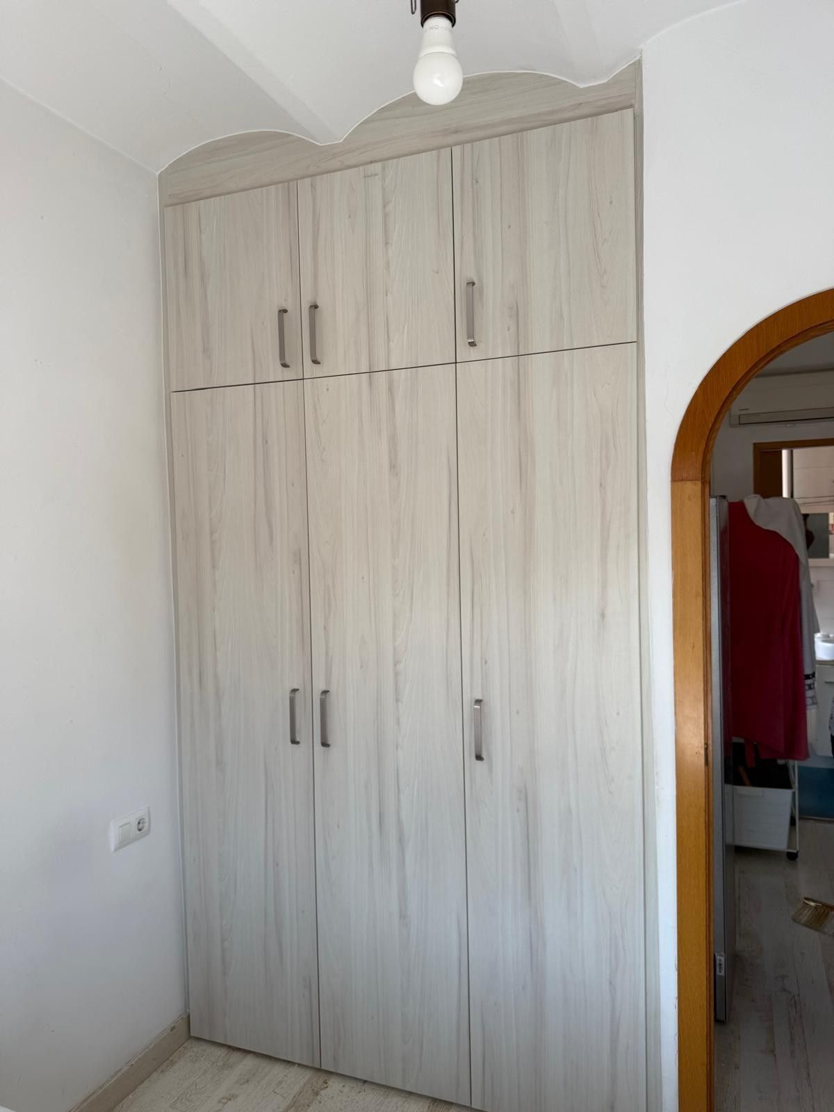 Armario de madera blanca con varias puertas en una pequeña habitación junto a una puerta arqueada.