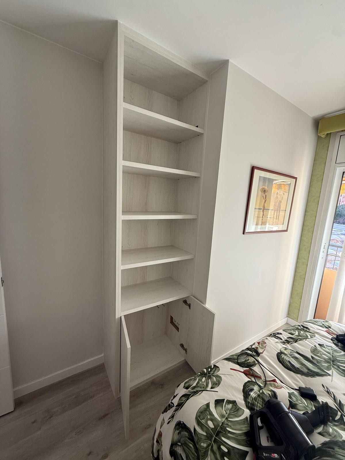 Estanterías blancas empotradas en un pequeño dormitorio junto a la cama, con un cuadro enmarcado en la pared.