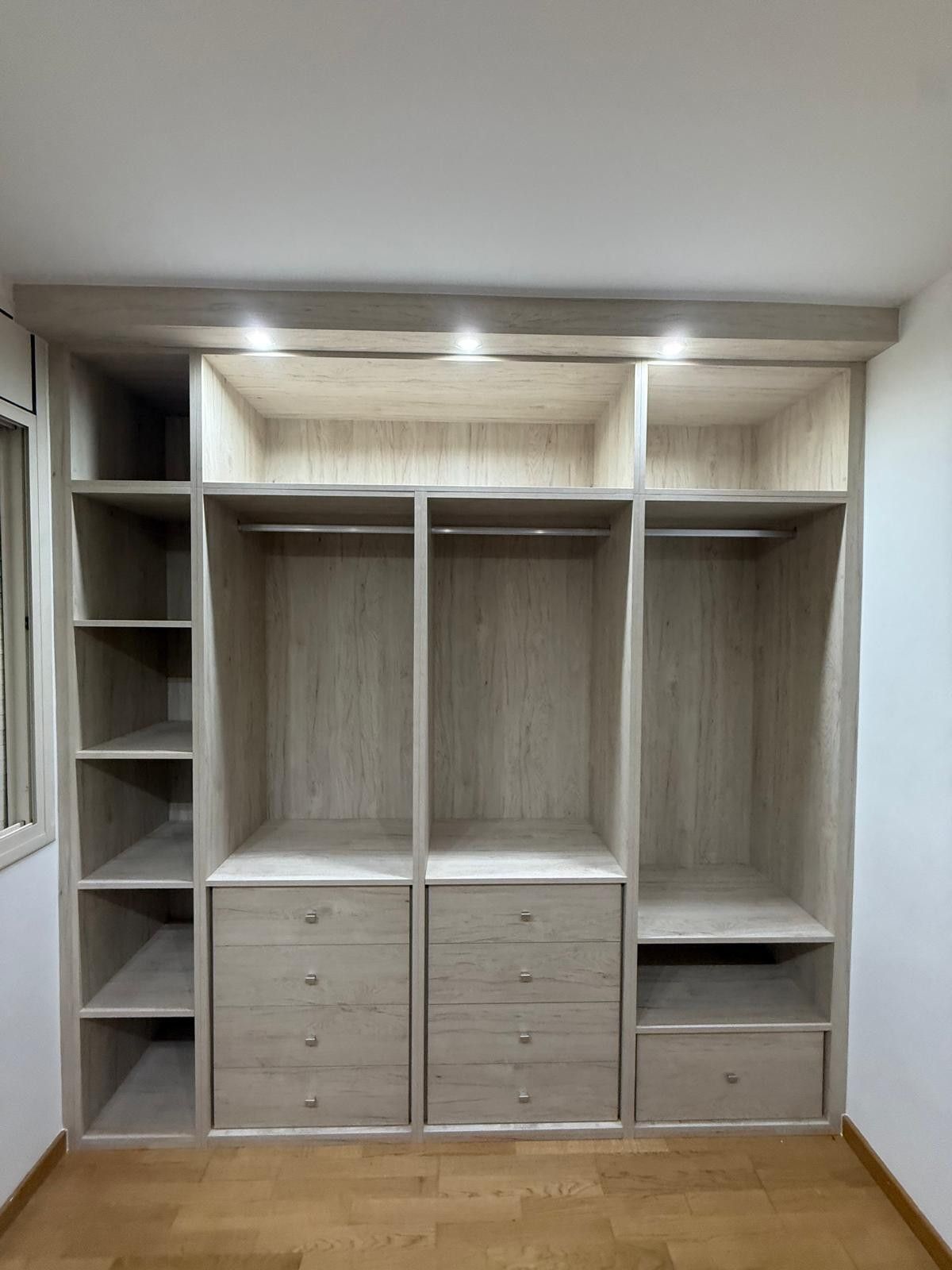 Armario empotrado de madera clara con estantes, cajones y espacio para colgar ropa, adosado a una pared blanca.