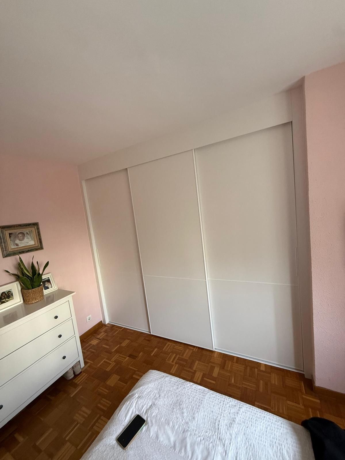 Dormitorio rosa con armarios blancos con puertas correderas, cómoda y cama con mando a distancia.
