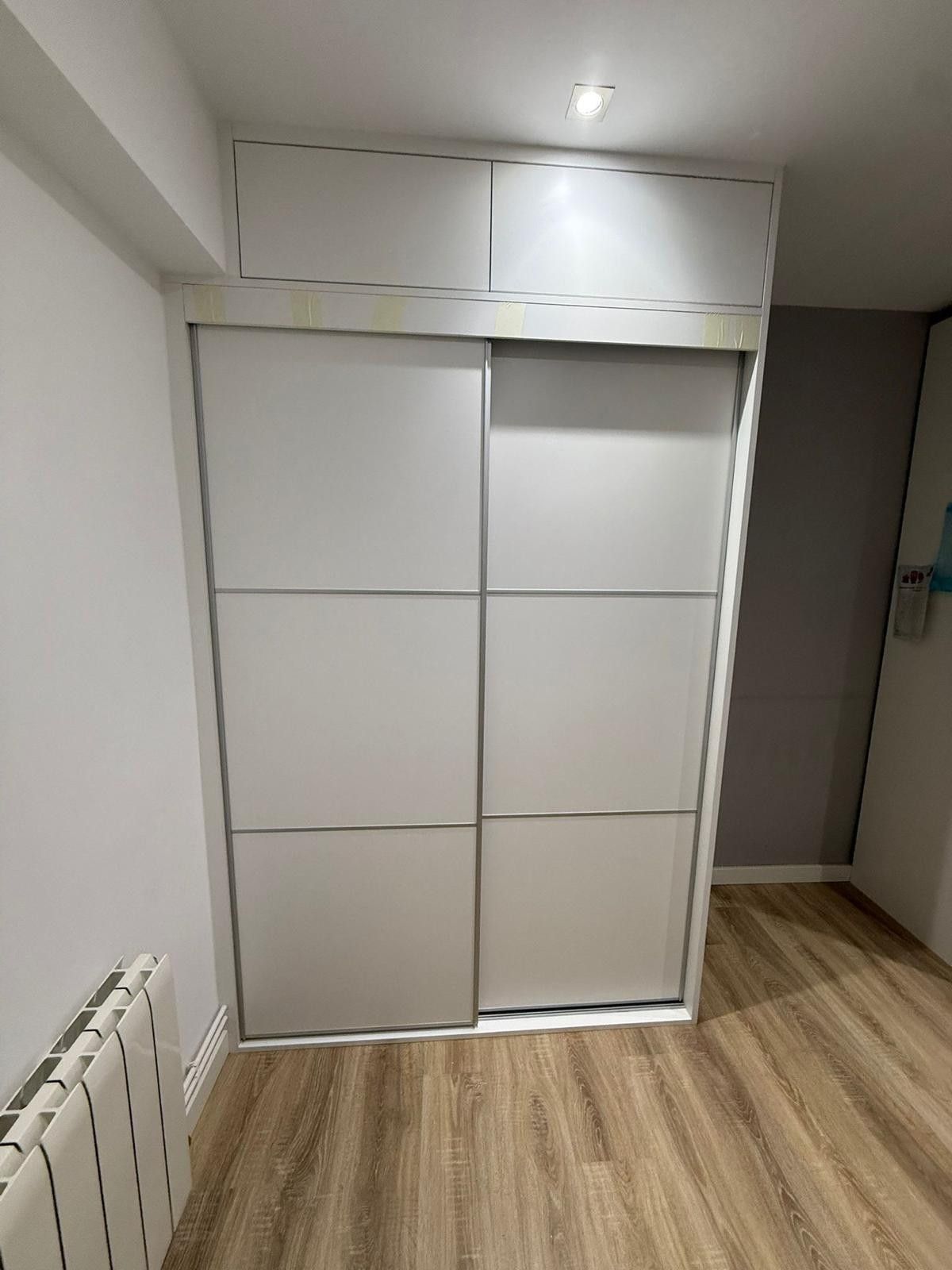 Puertas correderas blancas de armario en una habitación pequeña con suelo de madera clara y un radiador a la izquierda.
