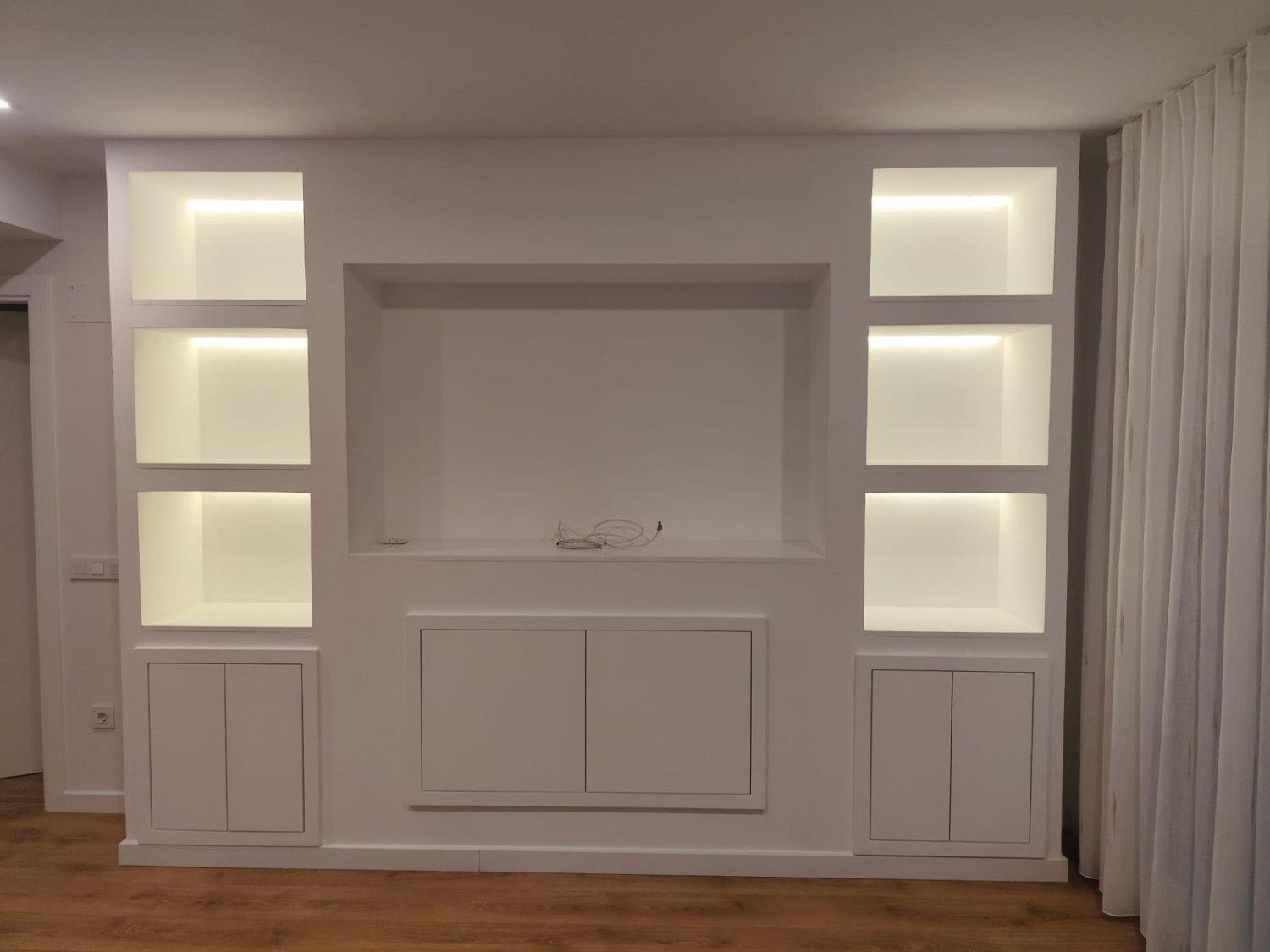 Mueble empotrado blanco con estantes iluminados y asiento junto a la ventana en una habitación de tonos neutros.