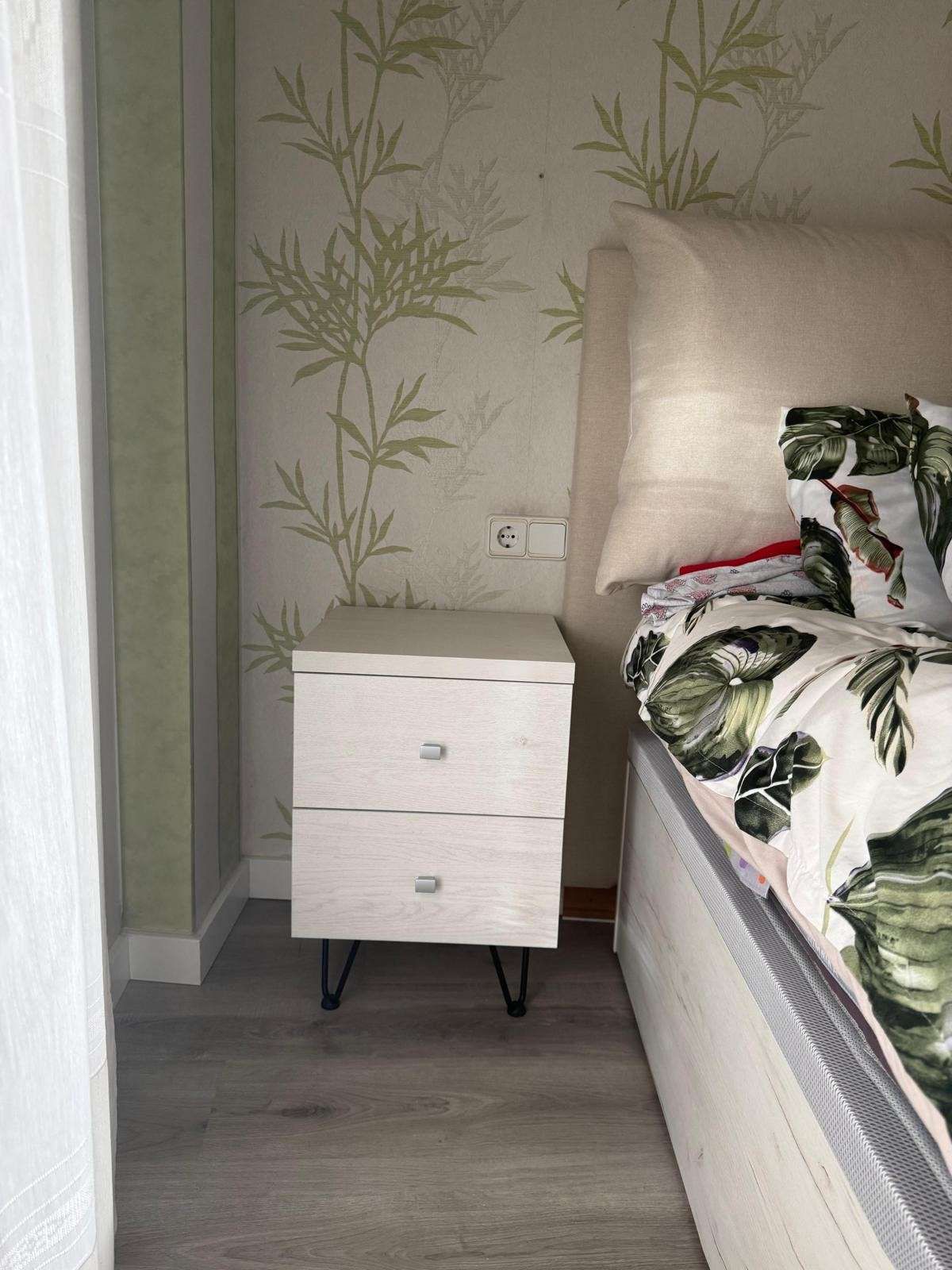 Mesita de noche pequeña de color blanco junto a la cama en un dormitorio con papel pintado con estampado de bambú.