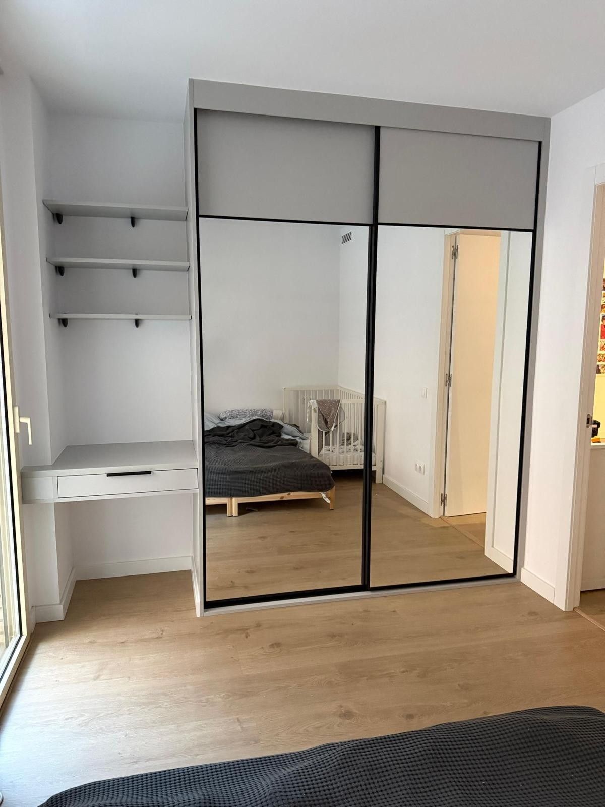 Habitación pequeña con armario con espejo, escritorio blanco, estantes de pared y suelo de madera.