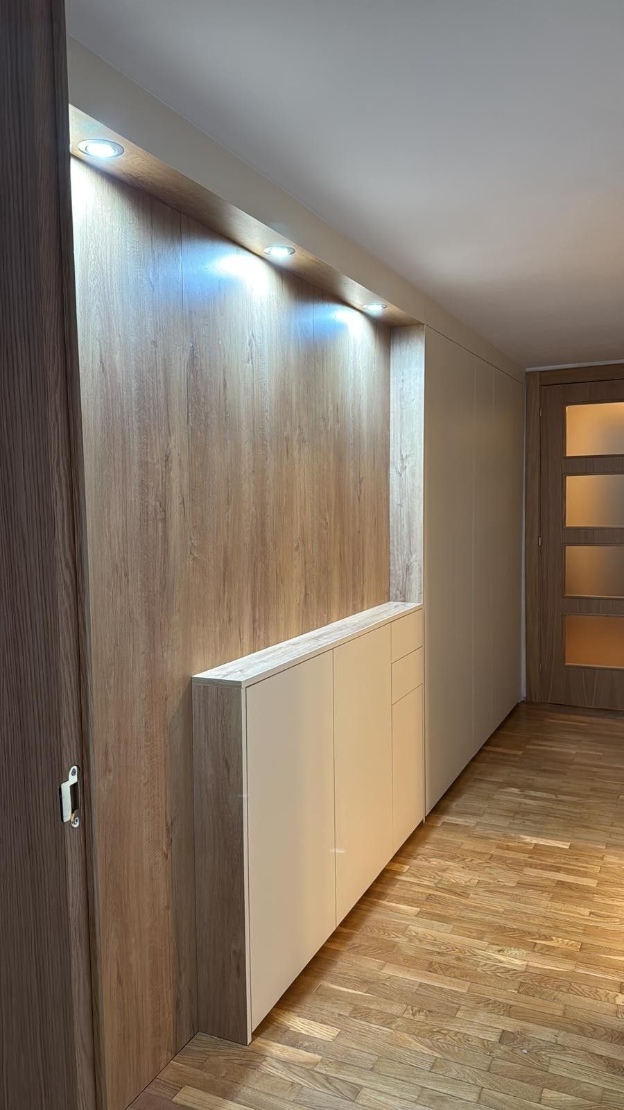Pasillo moderno con pared revestida de madera, luces empotradas, armario blanco y suelo de madera.