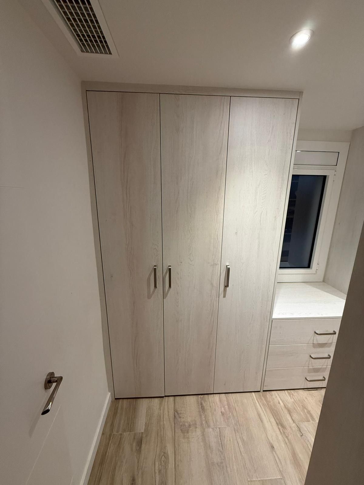 Habitación pequeña con armario blanco, escritorio con cajones, ventana y suelo de madera clara.