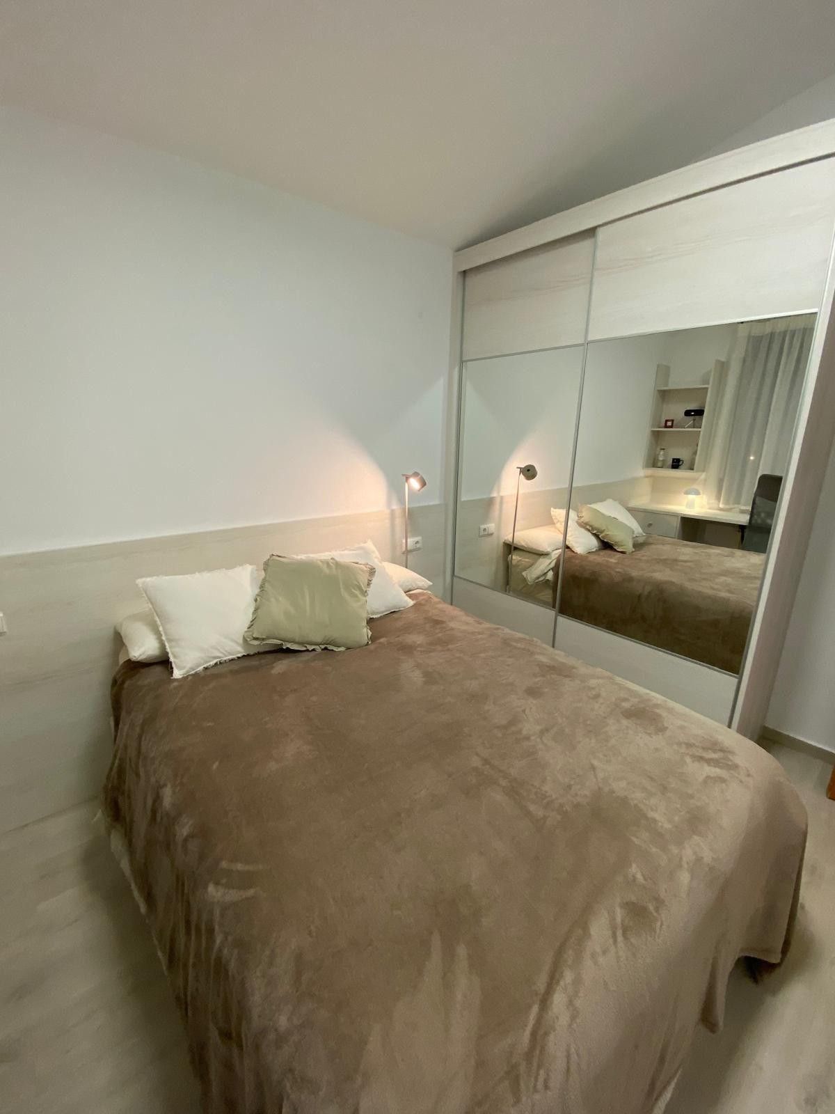 Habitación pequeña con cama, manta beige, almohadas blancas y puertas de armario con espejo.