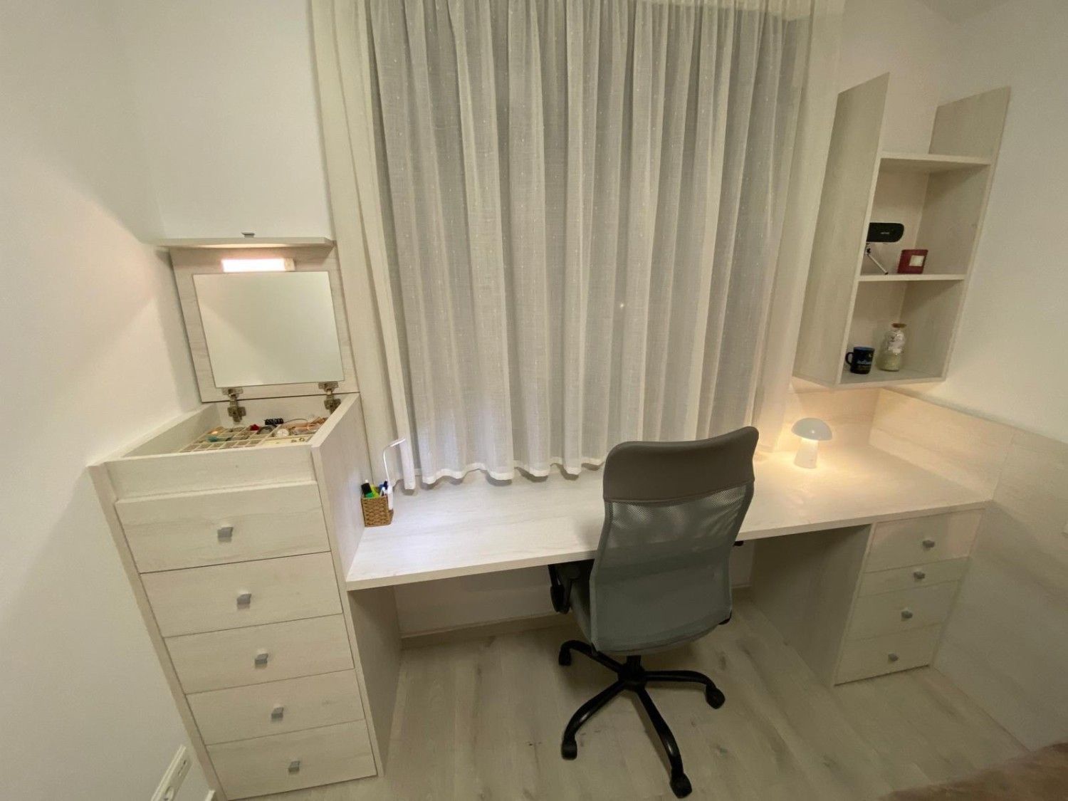 Escritorio blanco con silla, cajones, espejo, cortinas y pequeños estantes en una habitación luminosa.