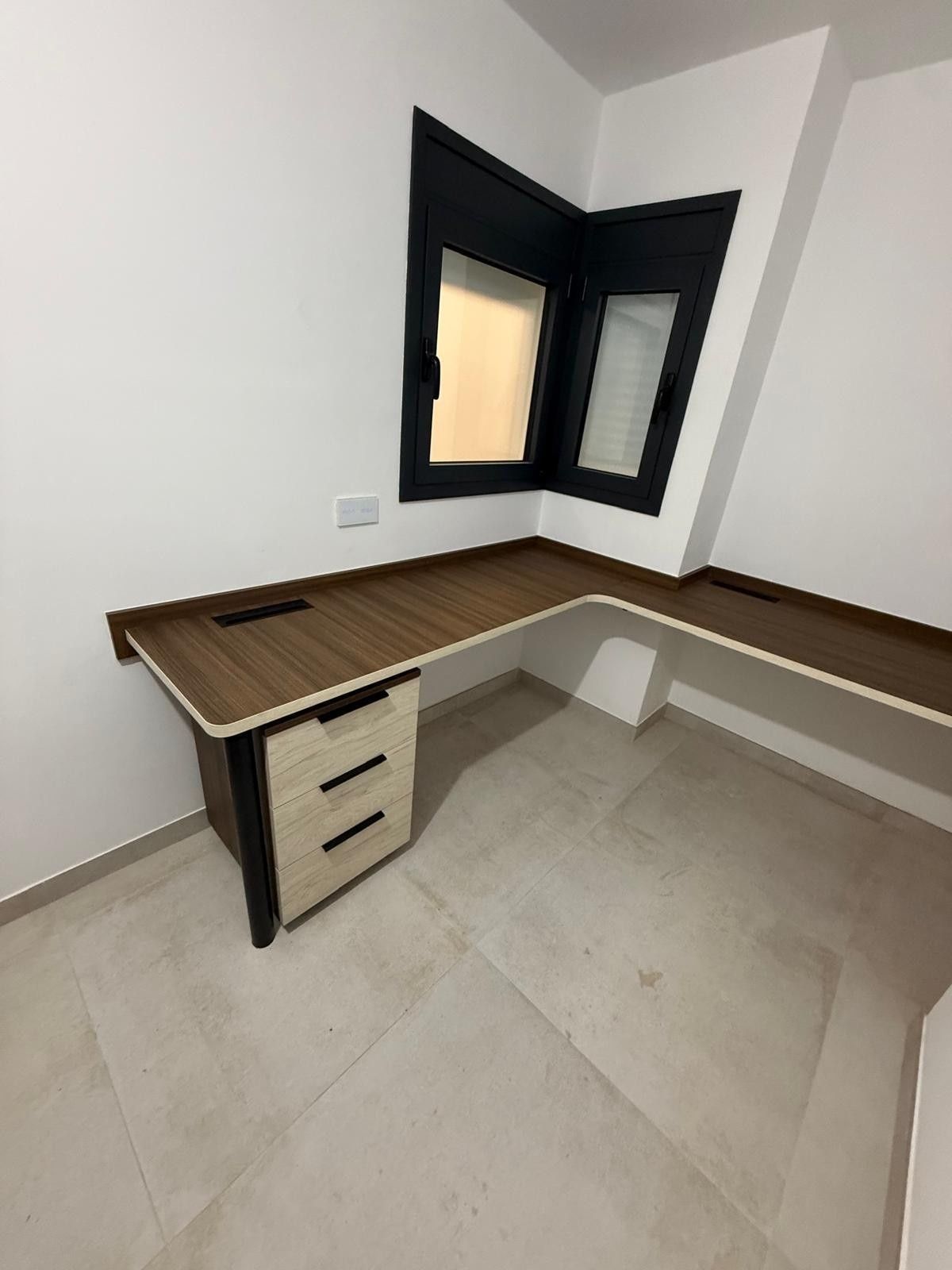 Escritorio esquinero vacío con tablero de madera, detalles en negro y cajones en una pequeña habitación blanca con ventana.