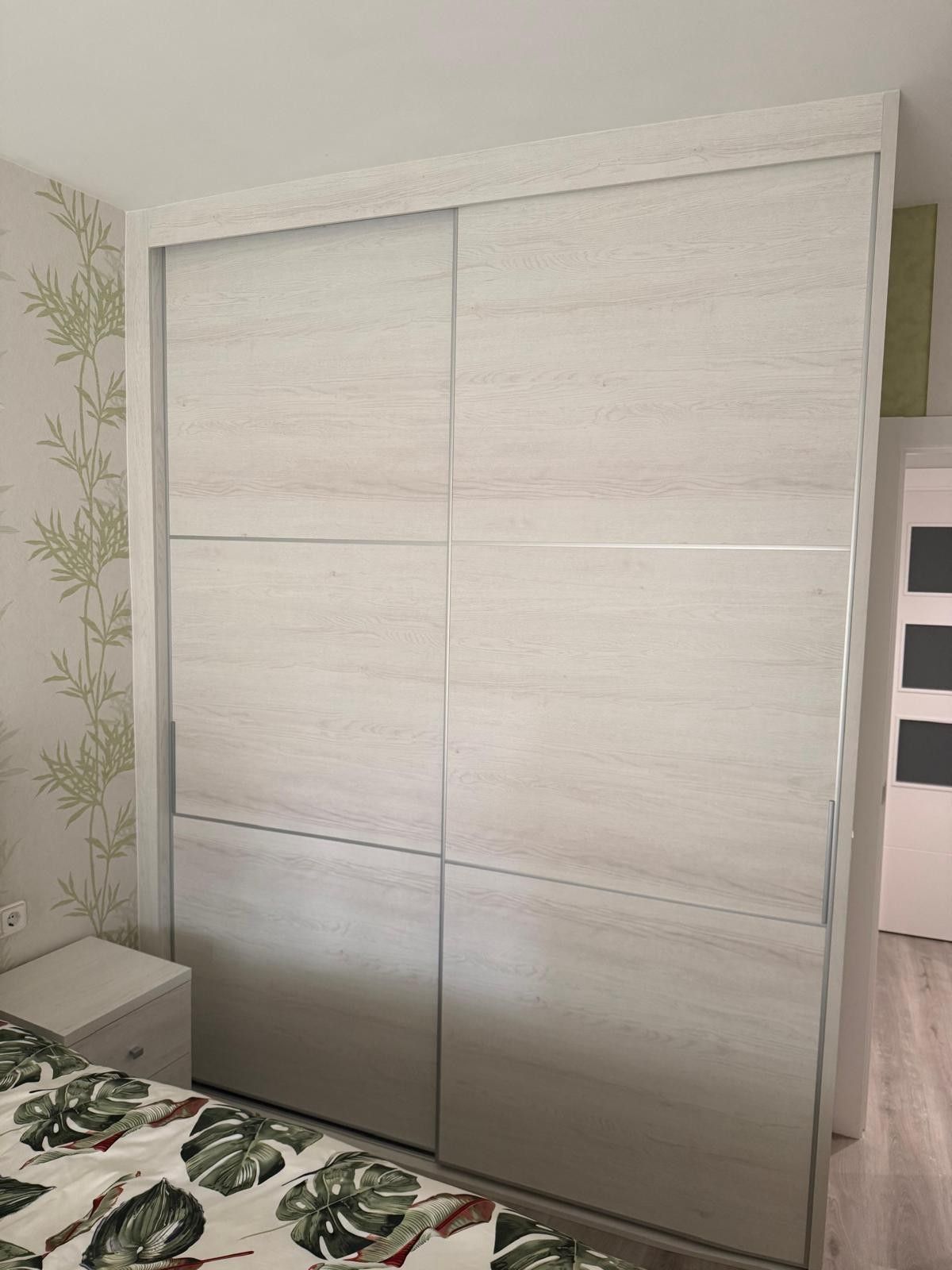 Puertas correderas blancas en un armario de dormitorio con papel pintado claro y ropa de cama floral.