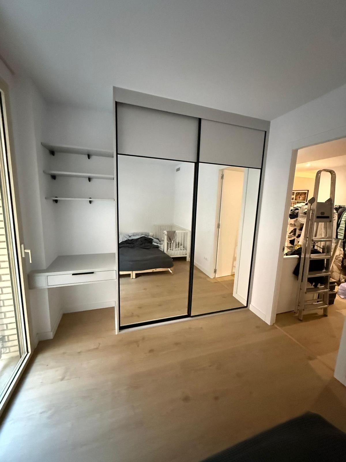 Habitación pequeña con armario con espejos, escritorio empotrado, estanterías y suelo de madera clara.