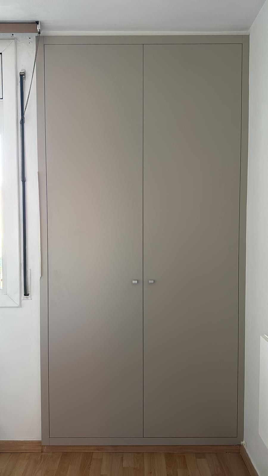 Armario beige de dos puertas contra una pared blanca, con una ventana a la izquierda y suelo de madera.