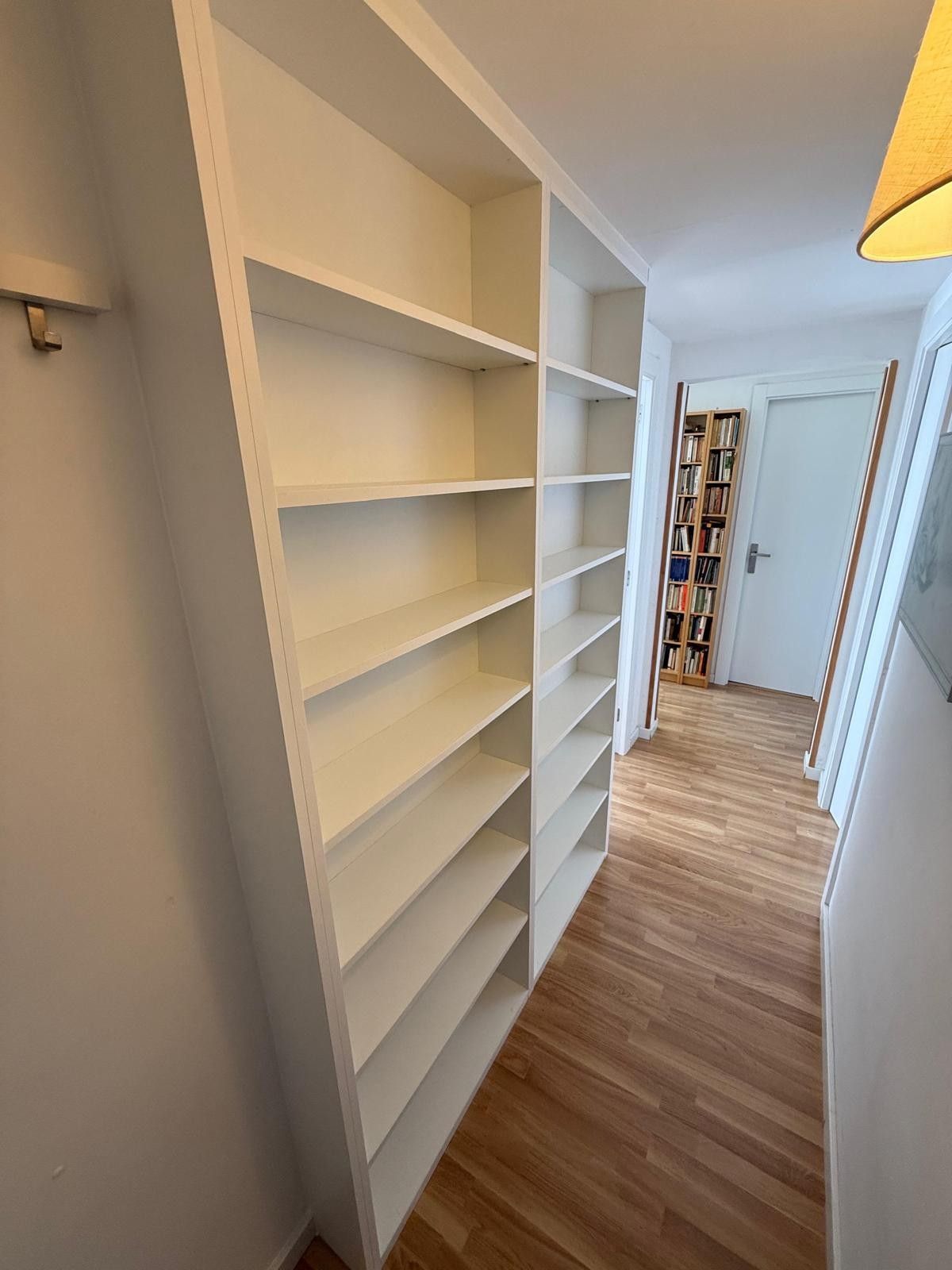 Pasillo estrecho con estanterías blancas empotradas y suelo de madera que conduce a una puerta.