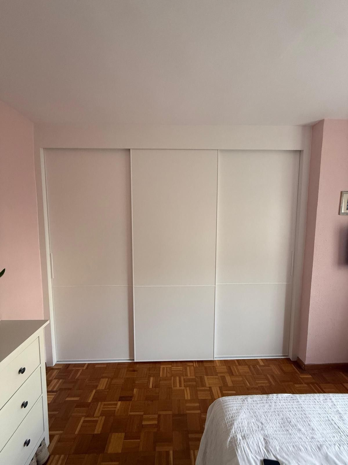 Habitación pequeña de color rosa con puertas de armario blancas, suelo de parqué, cómoda y parte de una cama.