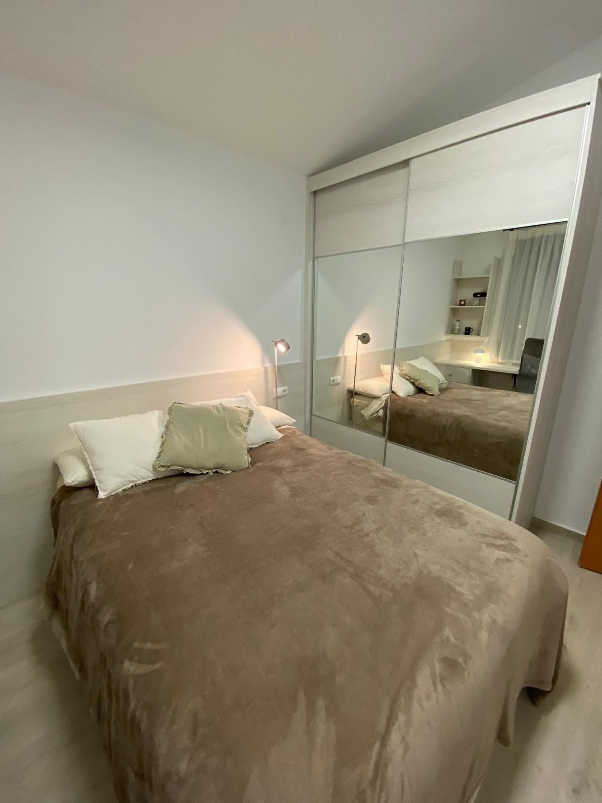 Dormitorio bien arreglado con colcha beige, armario con espejo, lámpara de noche y paredes blancas.