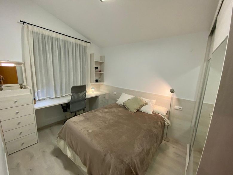 Habitación luminosa con cama sin hacer, escritorio junto a la ventana, cómoda y puertas de armario con espejo.