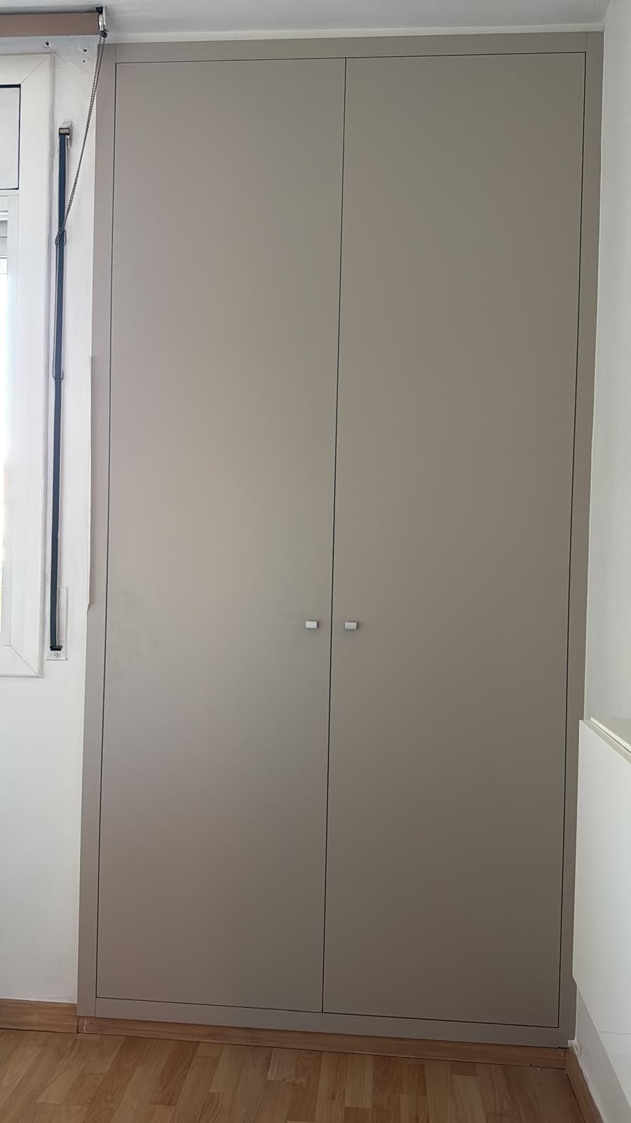 Armario alto de doble puerta de color beige situado entre las ventanas de una habitación pequeña con suelo de madera.