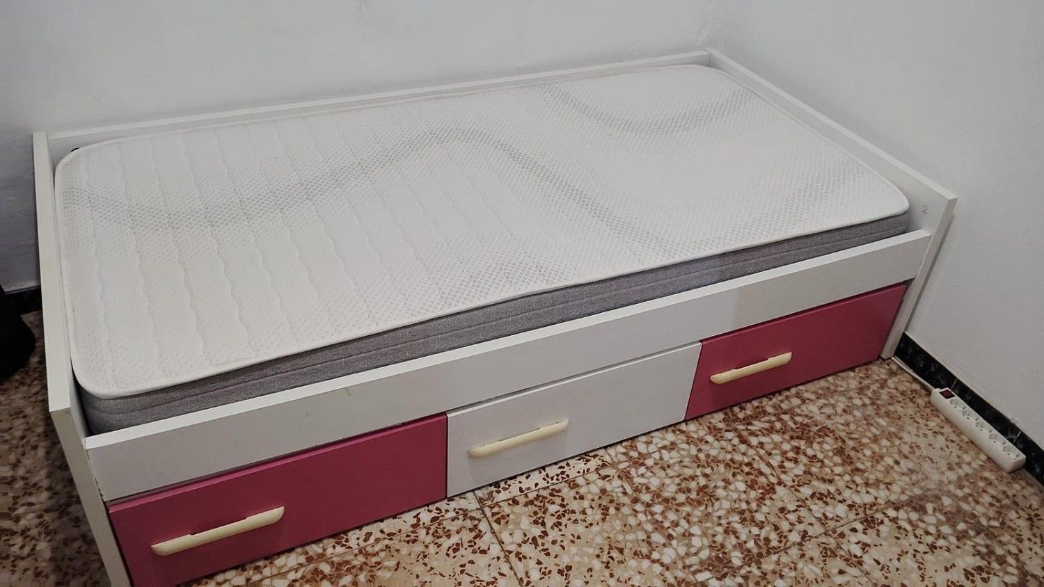 Colchón blanco sobre una cama plegable gris y rosa en un rincón alfombrado del dormitorio.