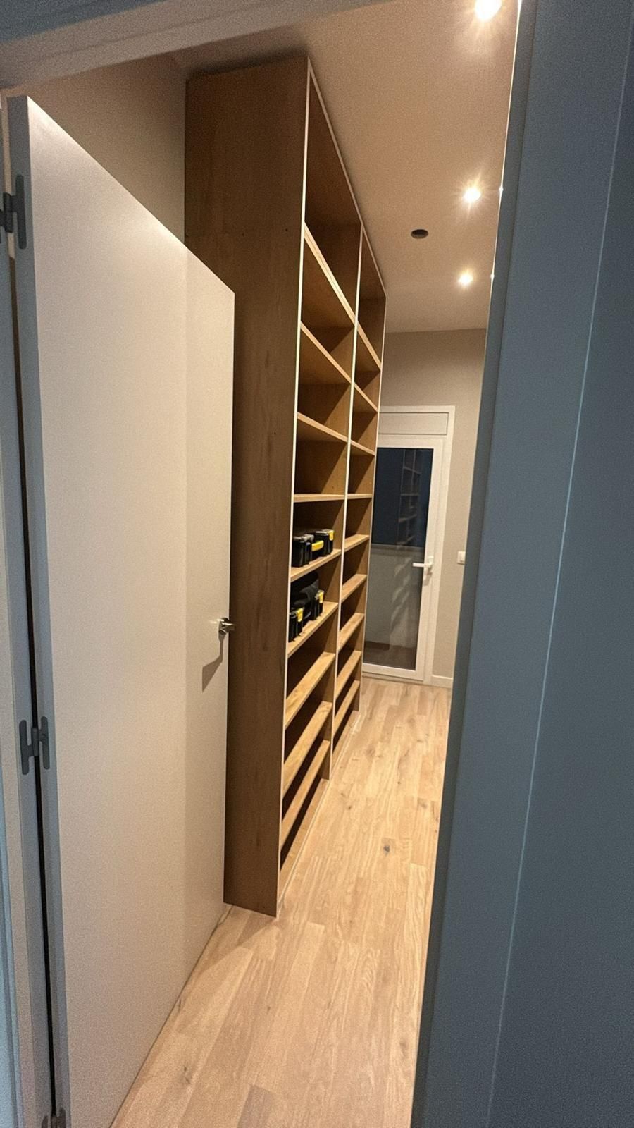 Pasillo estrecho con suelo de madera clara, paredes blancas, estanterías abiertas y una puerta al final.