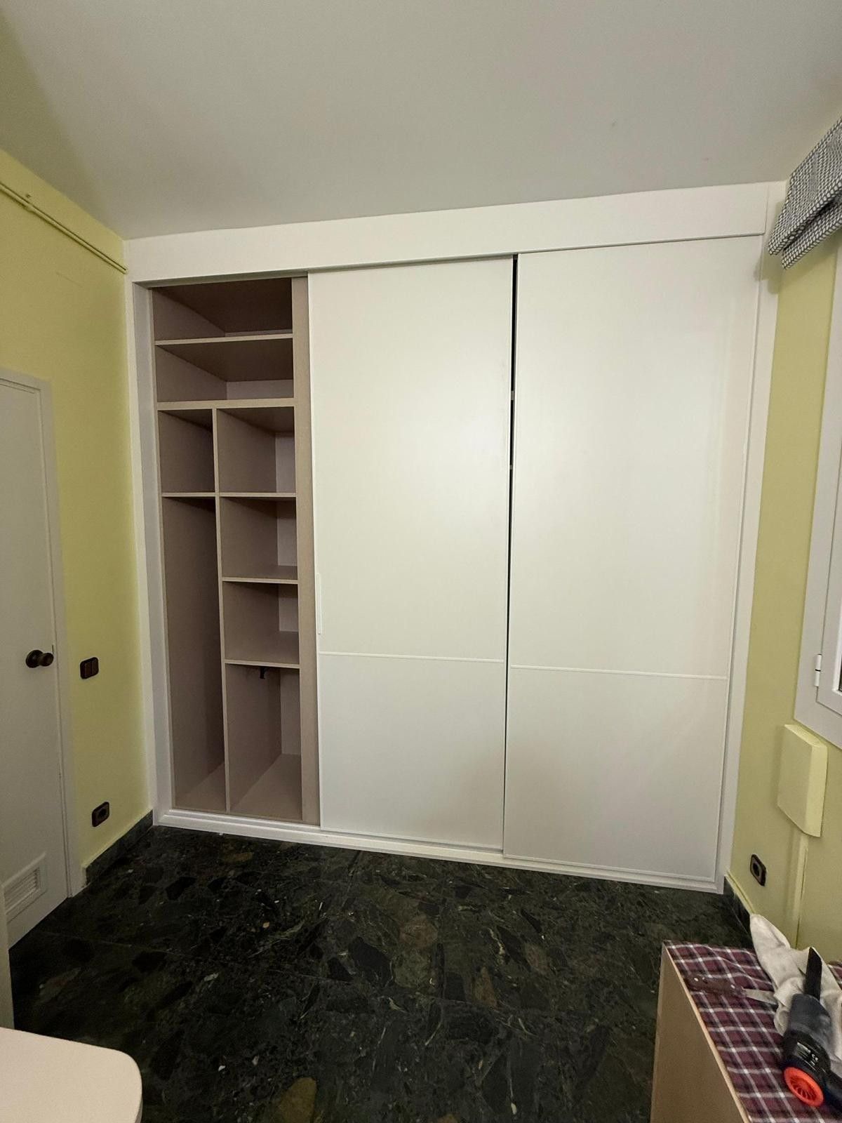 Habitación pequeña con armarios empotrados blancos y estanterías abiertas, paredes amarillas y suelo oscuro.