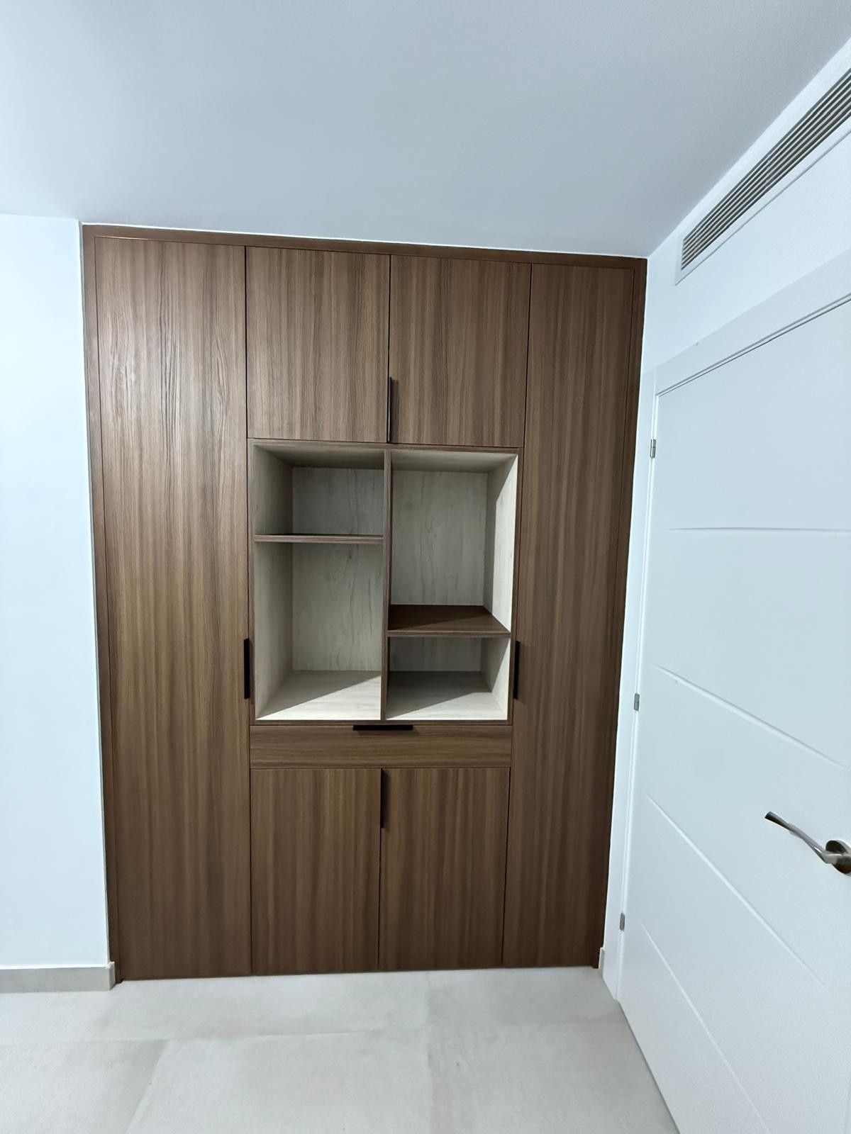 Un rincón con estanterías de madera empotradas en un pasillo blanco, junto a una puerta blanca.