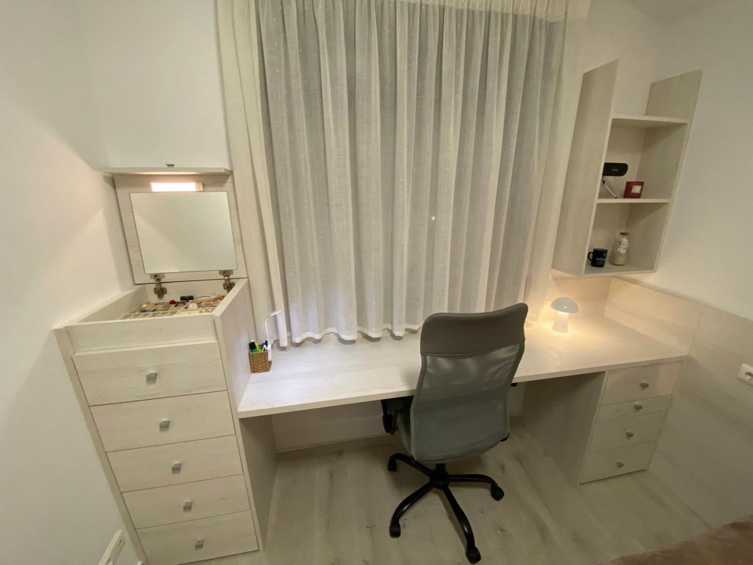 Rincón de escritorio blanco con espejo, silla, cajones y cortinas en una habitación luminosa.