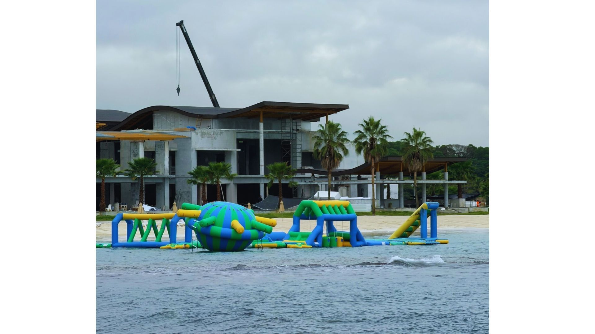 Water park obstacle course on beach in front of a building with crane; cloudy sky.
