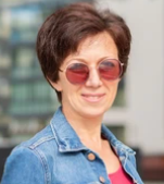 Woman in sunglasses, coat, touching face, inside an arched building.