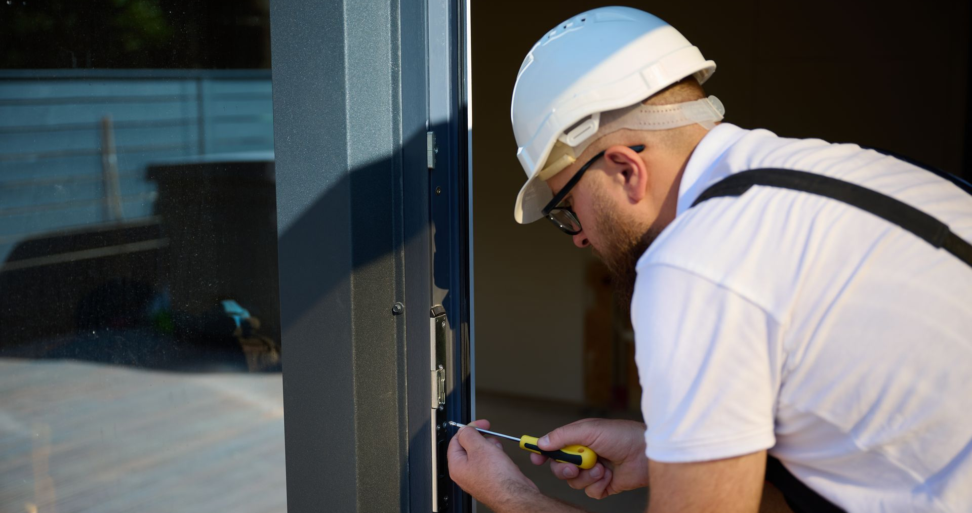 Une personne portant un casque de chantier répare un cadre de porte gris foncé à l'aide d'un tournevis.