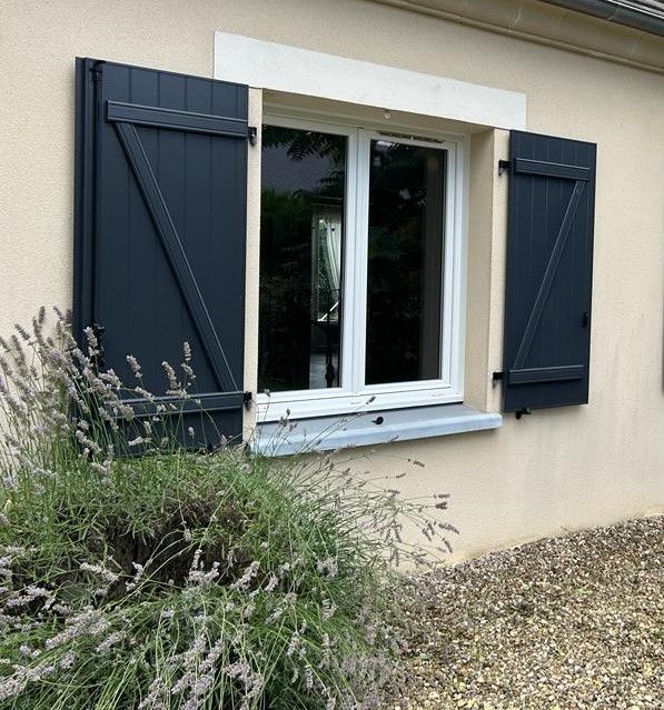 Des volets verts encadrent des portes-fenêtres blanches sur une maison blanche au toit de tuiles brunes.