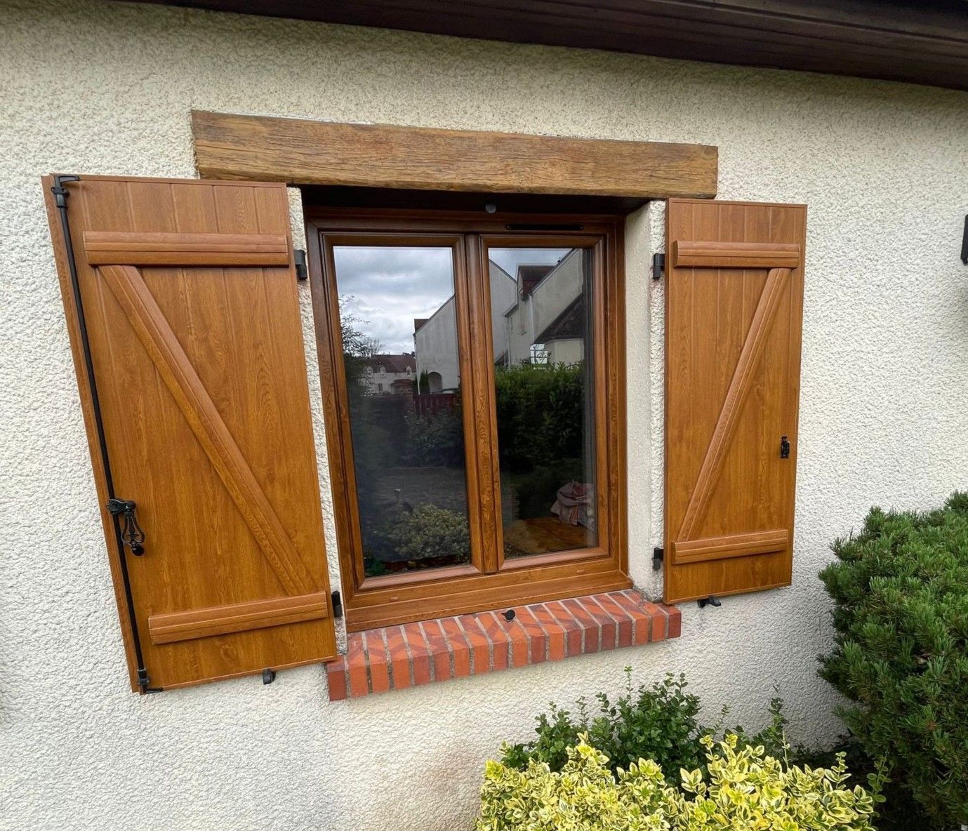 Fenêtre avec volets bruns, encadrement en bois et appui de fenêtre en briques sur un mur en stuc clair.