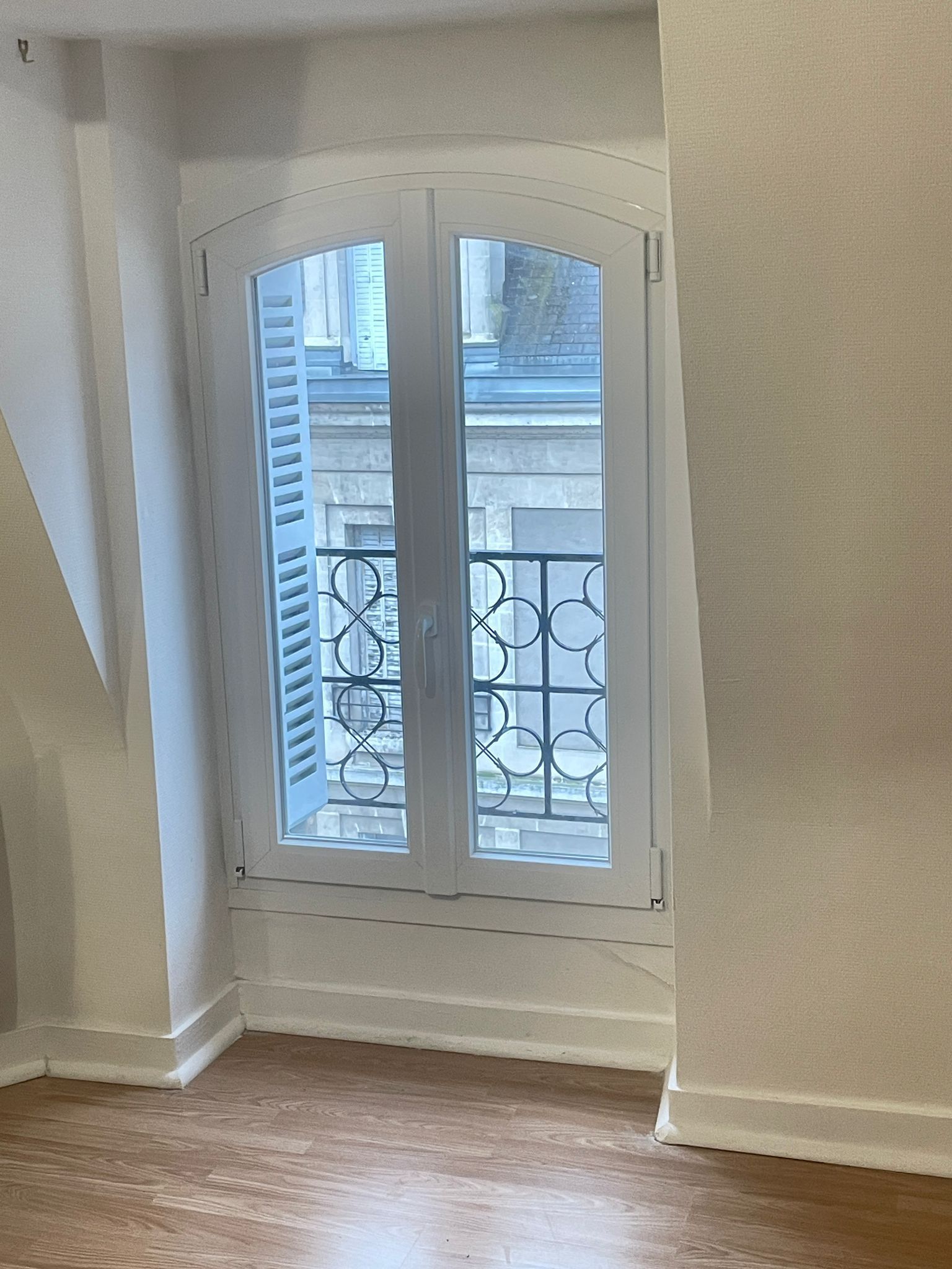 Dans une pièce au parquet en bois dur, une haute fenêtre cintrée aux encadrements blancs et un radiateur sont adossés à un mur blanc.