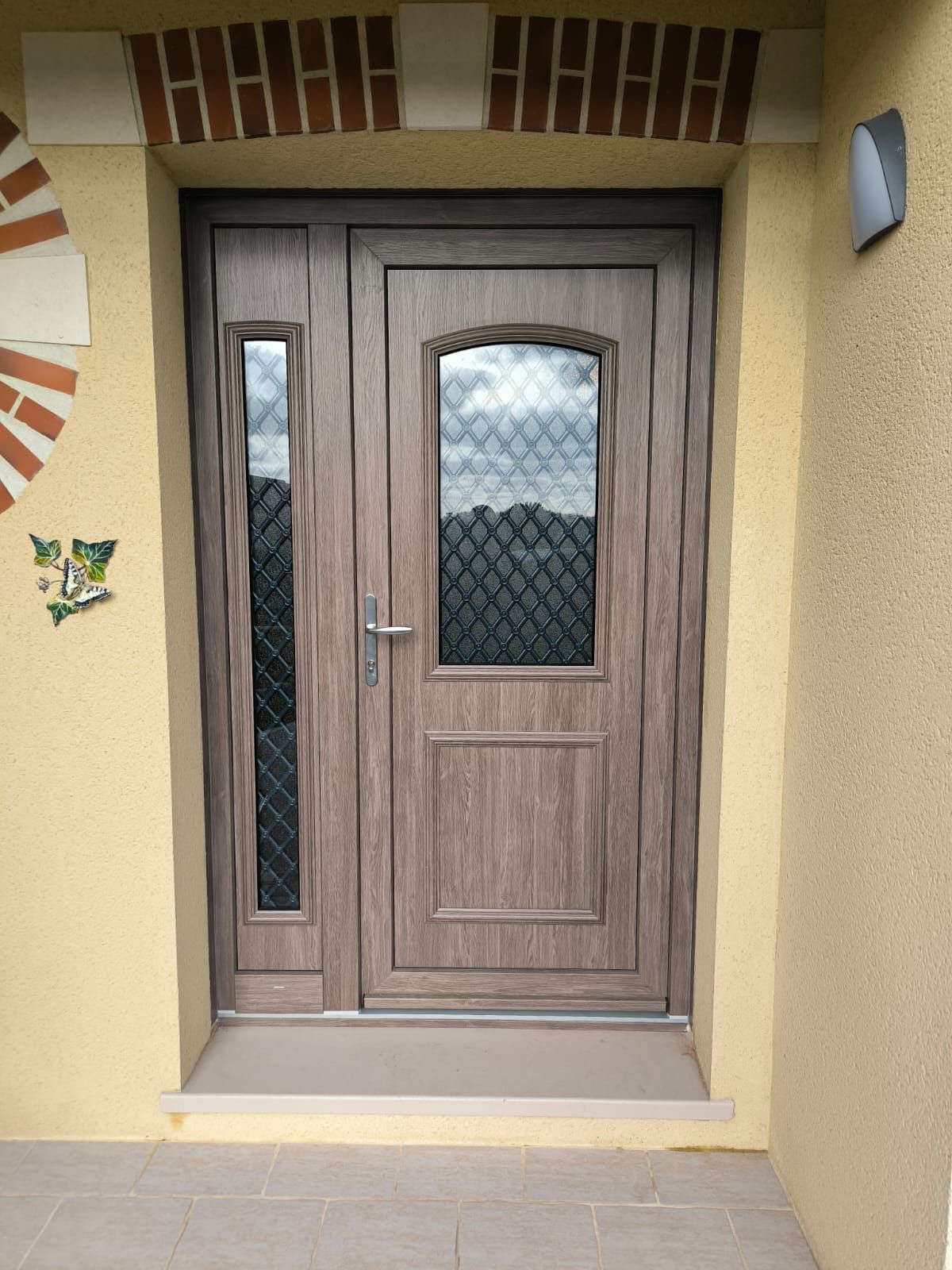 Une porte d'entrée avec un panneau latéral, toutes deux dotées de vitrages décoratifs, dans un cadre brun clair, encastrée dans un mur extérieur beige.